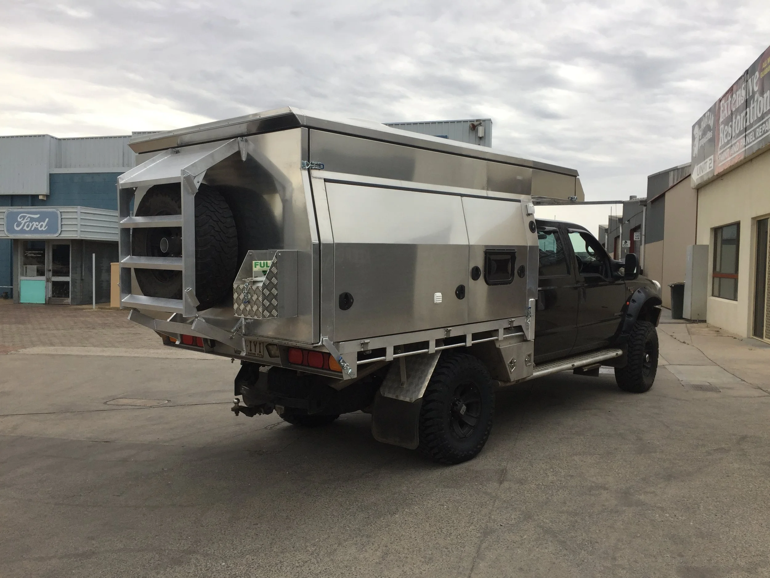 A black pickup truck with a large, silver, enclosed utility or custom service body attached to the bed, parked outdoors near a building. The utility body has a spare tire mounted at the rear and appears to be used for work purposes.