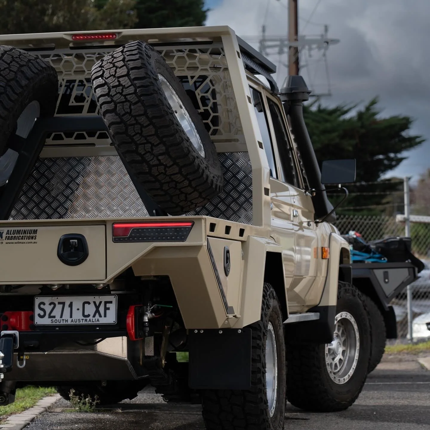 A beige 79 series Landcruiser with a custom tray and mounted spare tire in the back, parked outdoors on a cloudy day.