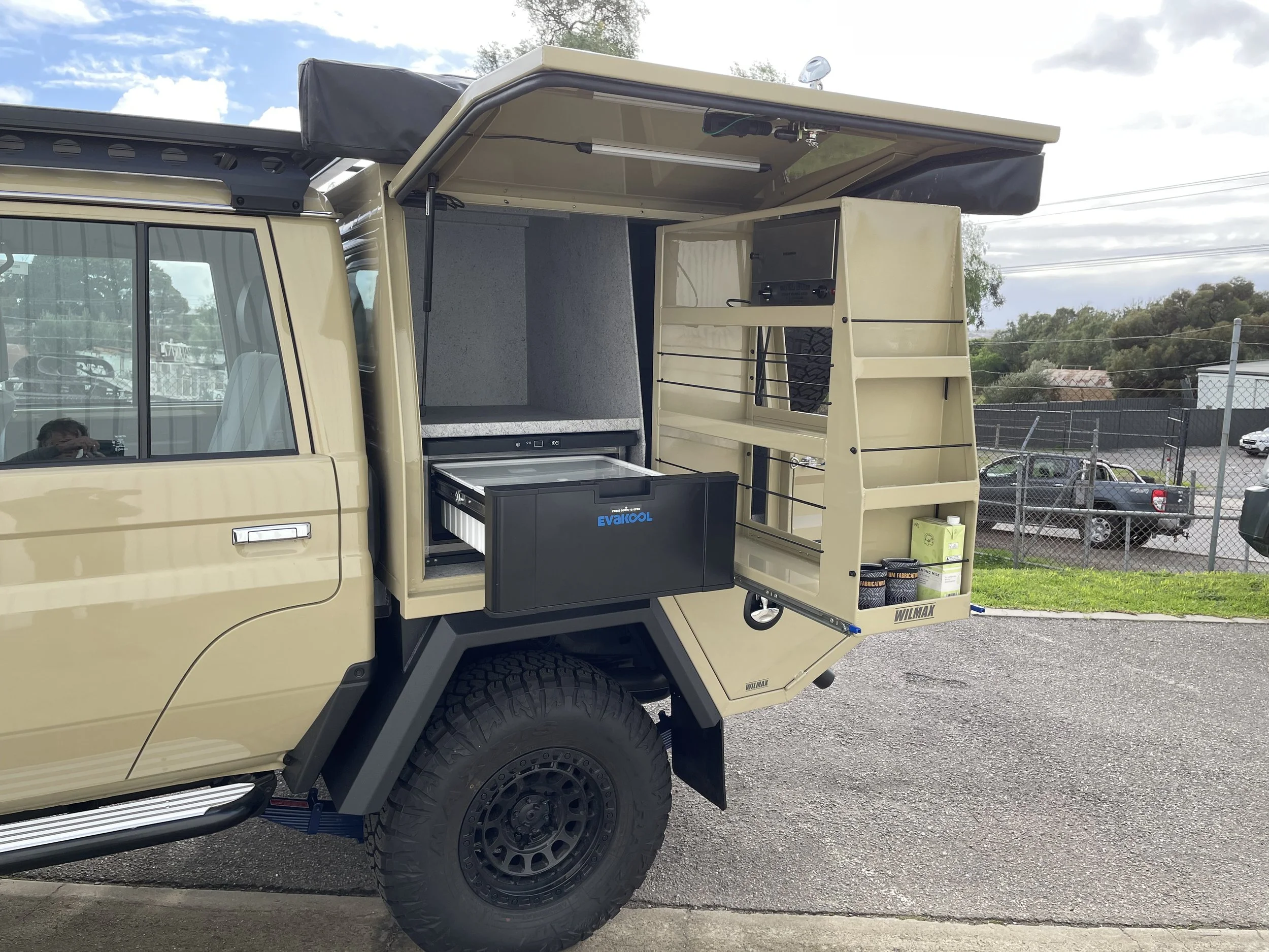 Side view of a beige off-road camper truck with open storage compartments at the back, parked on a gravel lot under a partly cloudy sky.