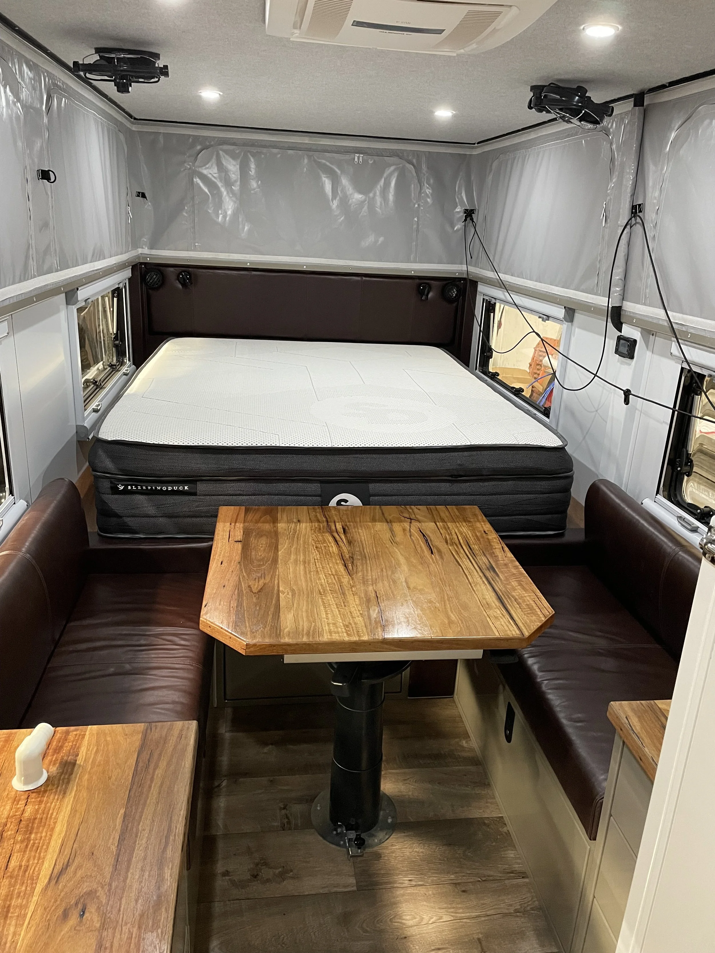 Interior of a small camper or RV with a bed, two windows, and a table with bench seating. The walls are covered in white insulation material and there are various electrical wires and components visible.