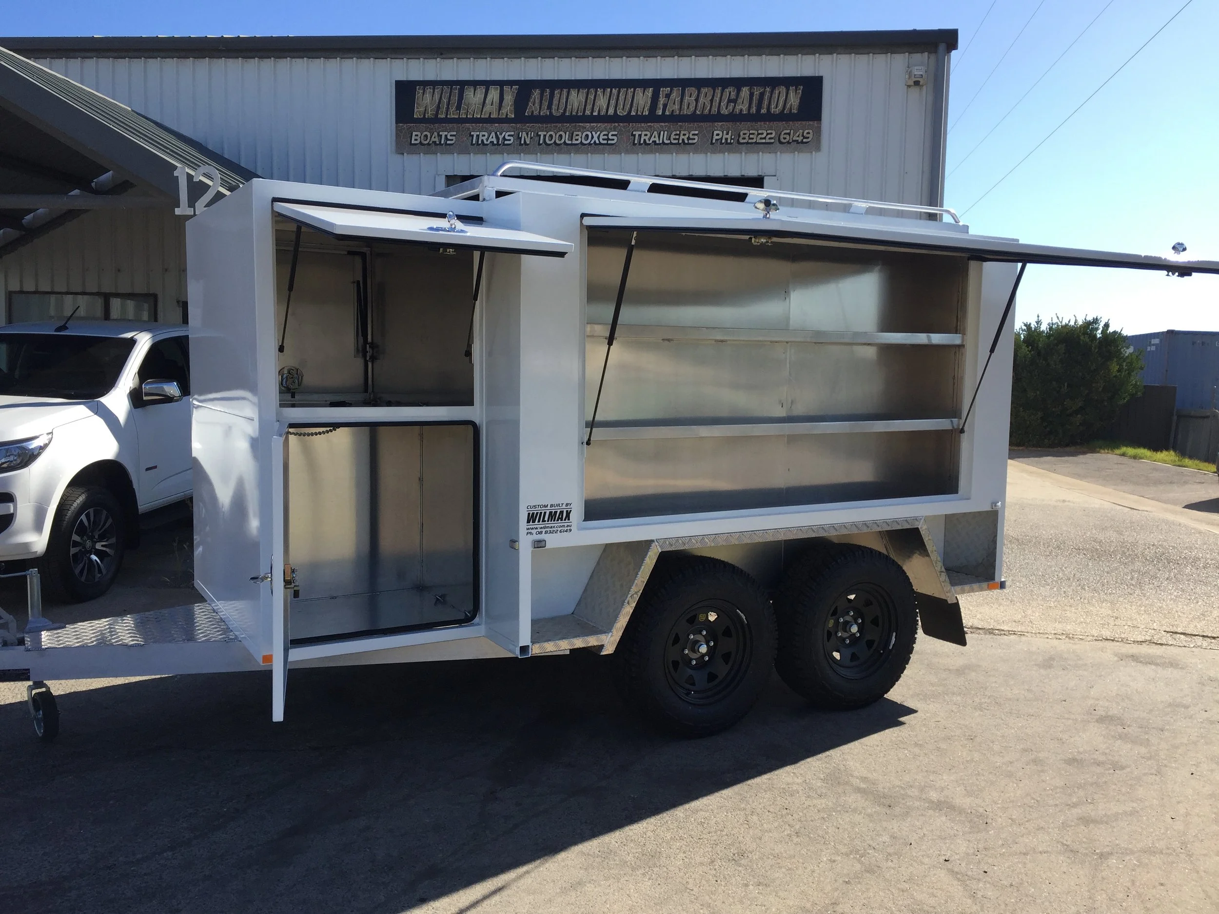 A white aluminum food and product trailer parked outside a building with a sign reading 'Wilmax Aluminum Fabrication.' The trailer has open side compartments and is positioned next to a white vehicle.
