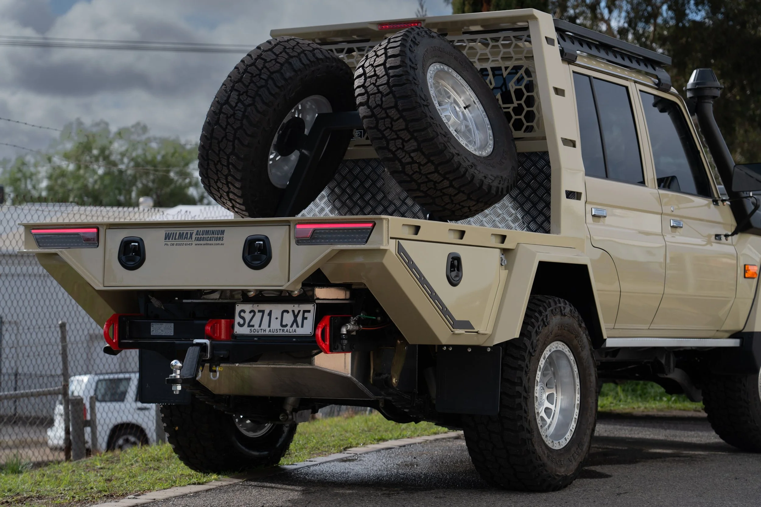 View of the back of A beige 79 series Landcruiser with a custom tray and mounted spare tire in the back,. The truck has a South Australia license plate, and the bed features toolboxes and utility compartments.