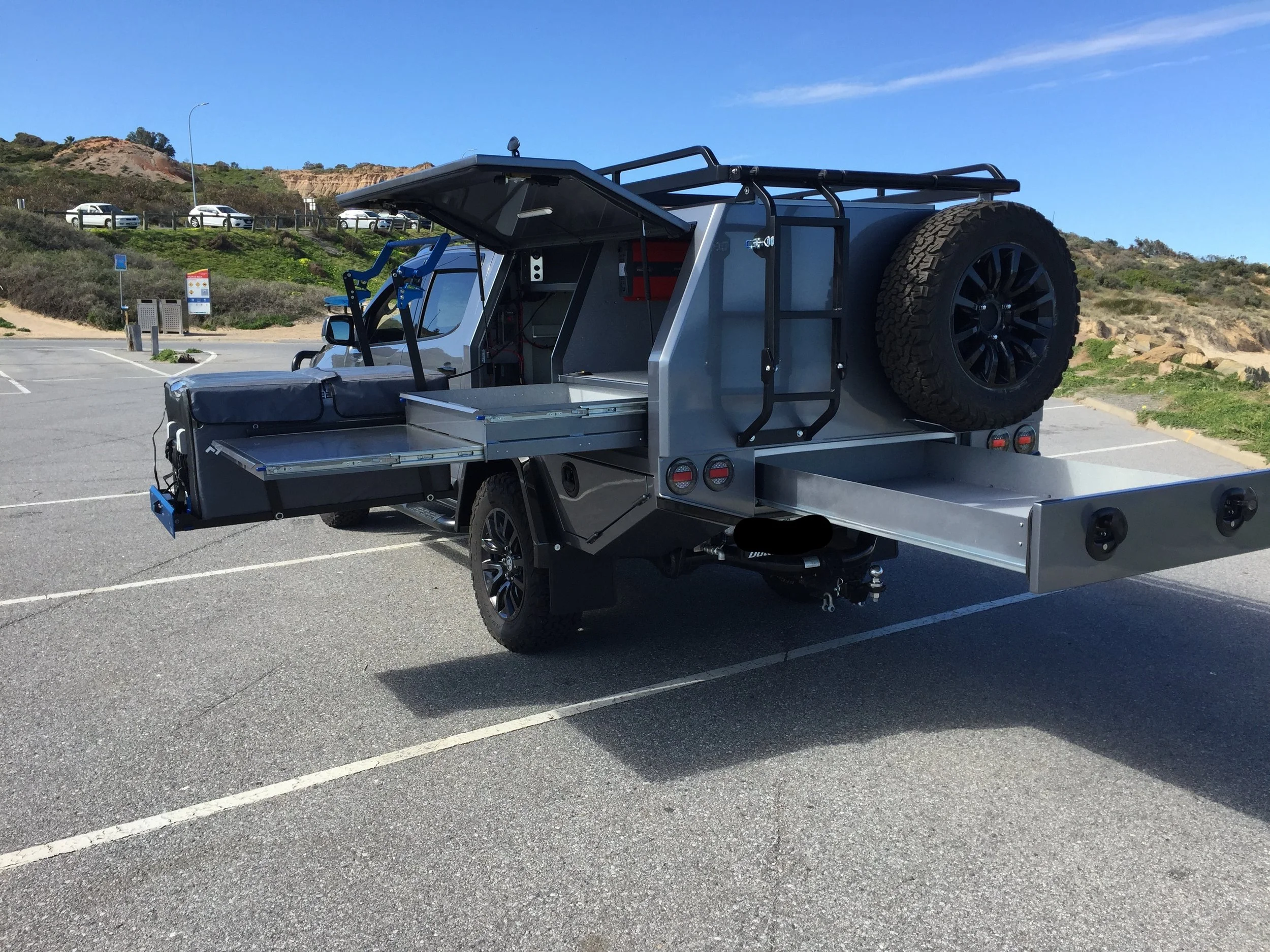 A silver utility truck with a large spare tire mounted on the back, parked in a lot on a clear day with a hillside and several parked cars in the background.