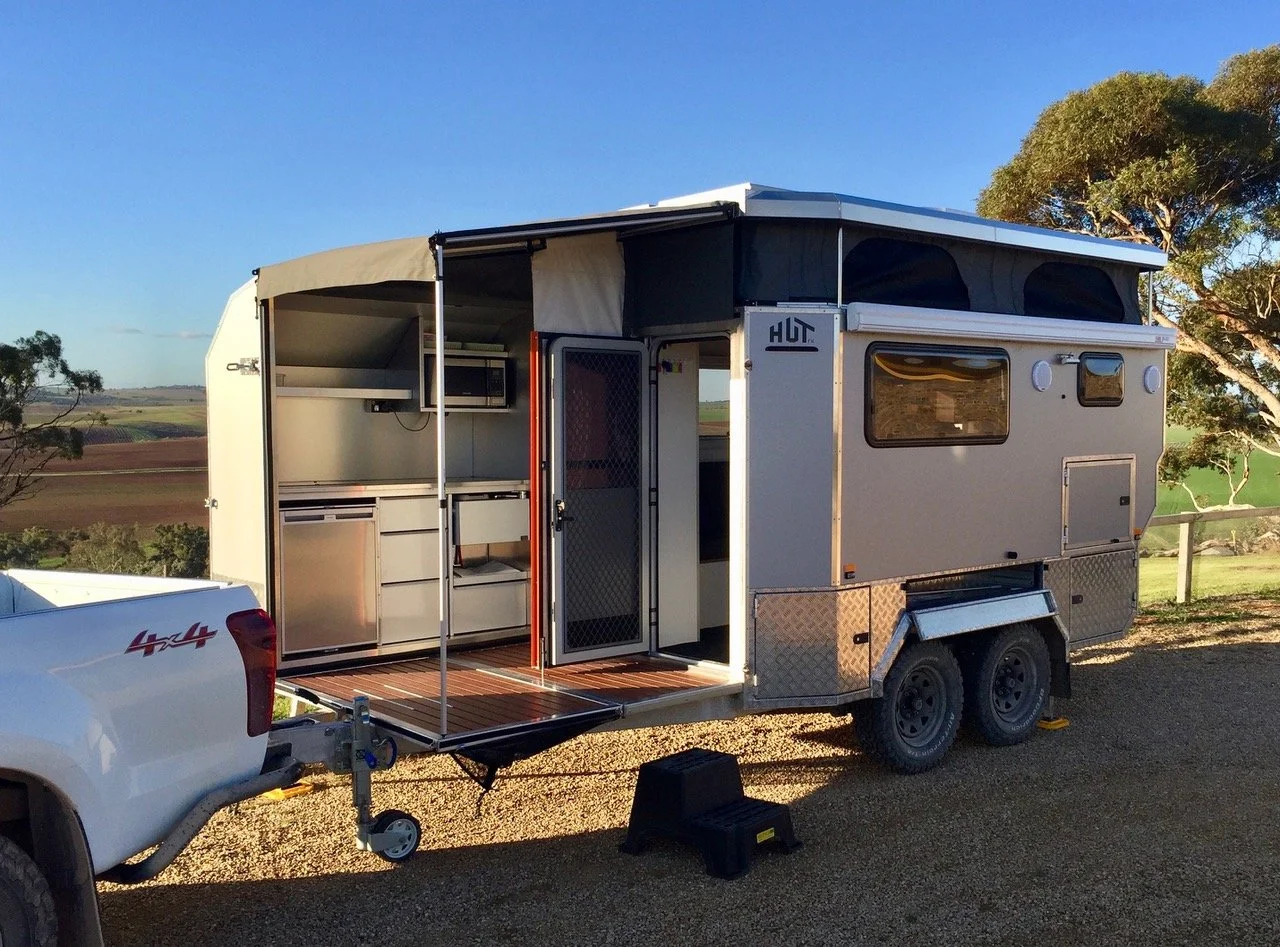Silver camping trailer hitched to a white pickup truck, parked outdoors on a gravel surface, with an open side showing interior kitchenette, under a clear blue sky with distant trees and fields.