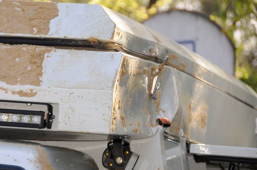 Close-up of a damaged white camper trailer with rust and dents, showing a damaged corner and a small ladybug on the metal surface.