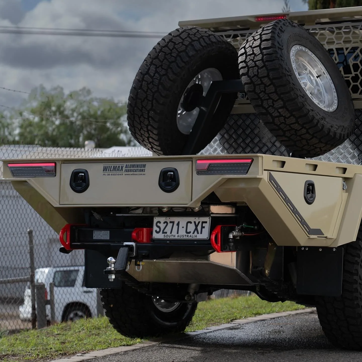View of the back of A beige 79 series Landcruiser with a custom tray and mounted spare tire in the back,. The truck has a South Australia license plate, and the bed features toolboxes and utility compartments.