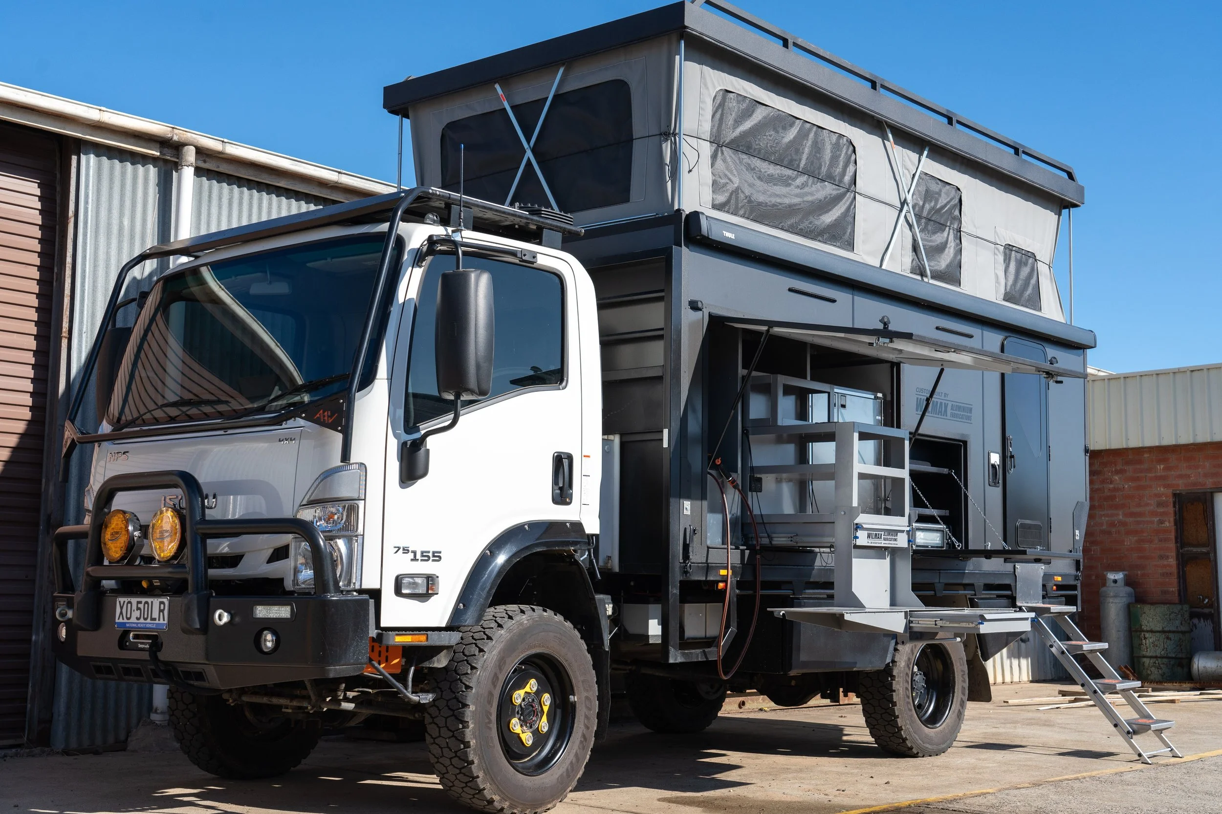 A white truck with a black bumper and dual yellow fog lights parked next to a building. The truck has a large, custom camper, possibly for mobile work or living space, with steps leading up to an entrance.