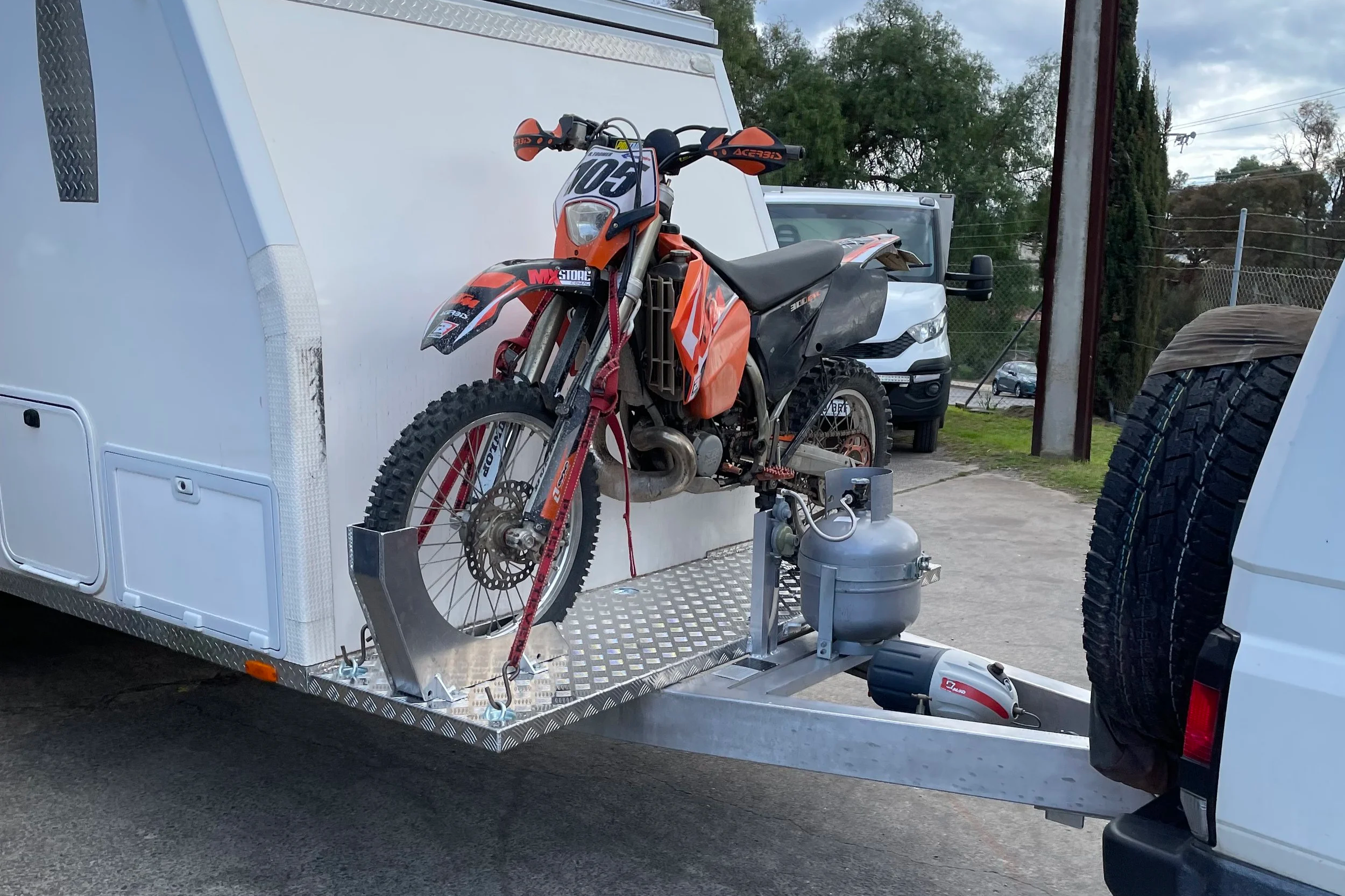 A dirt bike motorcycle mounted on a trailer with a propane tank attached, parked outside near trees and vehicles.