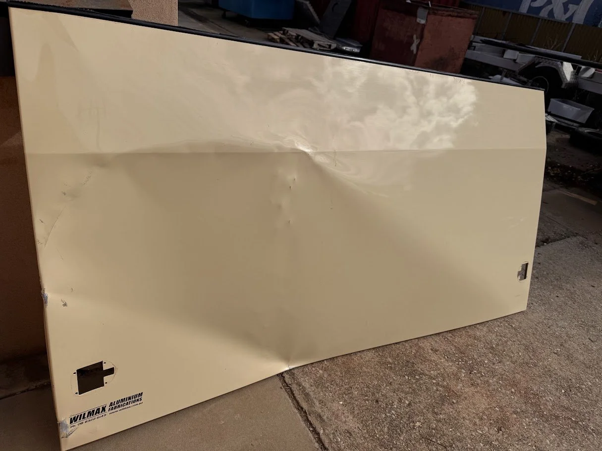 A large, beige, dented metal panel or sign leaning against a wall outdoors. It has a logo that reads 'Wilmax Aluminum Fabrications' and some cut-out sections.