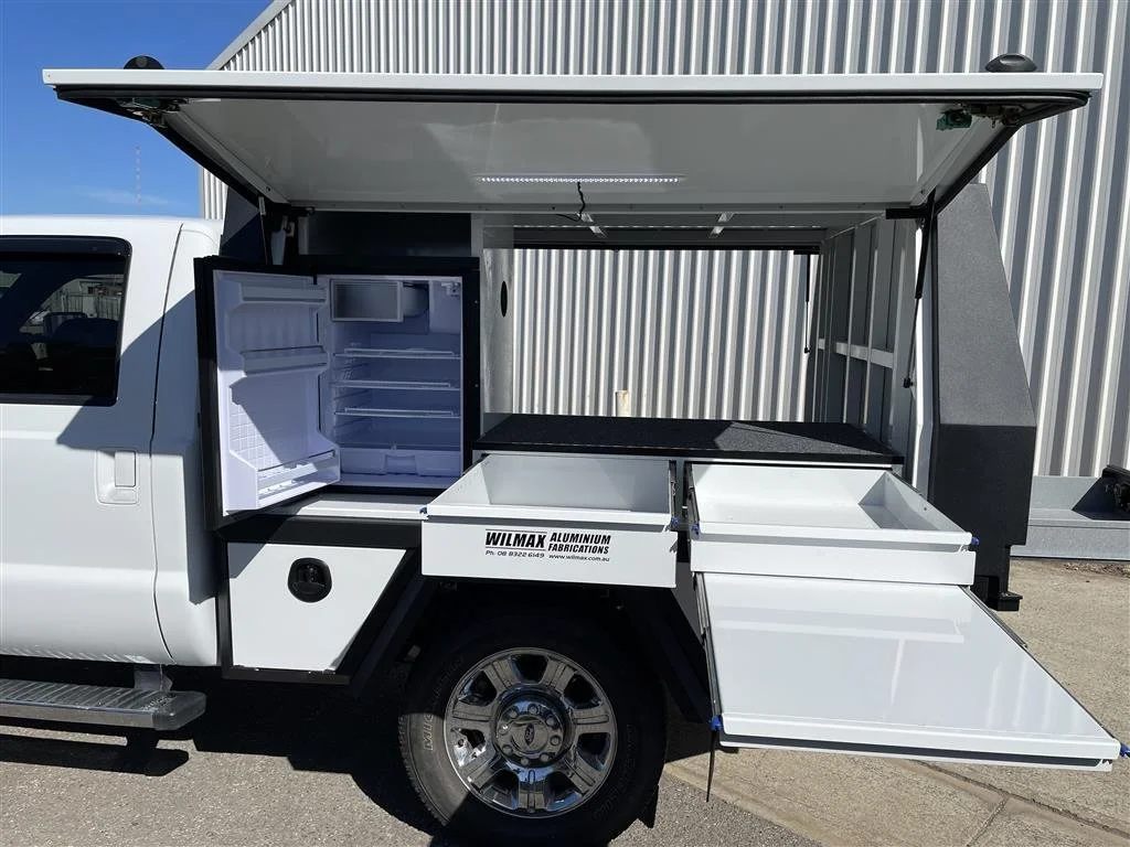 White utility truck with custom canopy, open storage compartments and a refrigerator in the back, parked in front of a metal building.