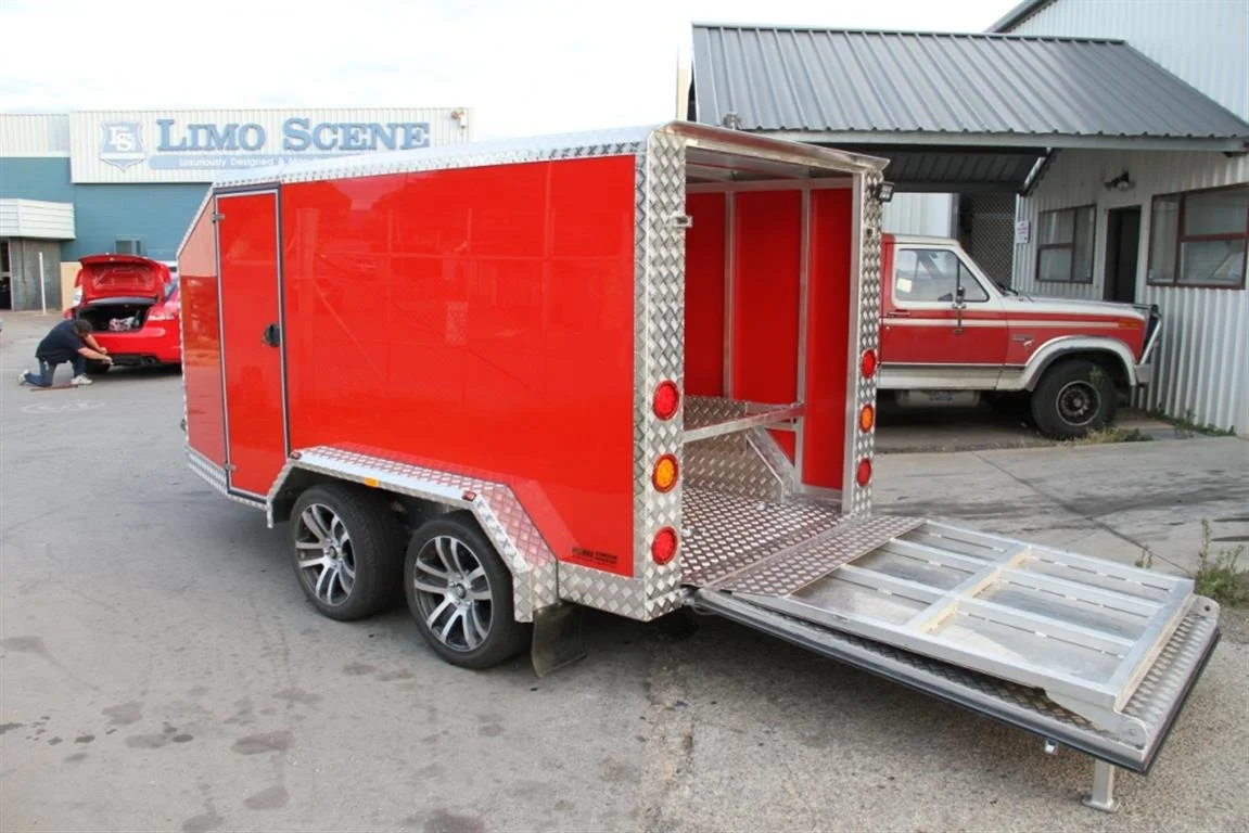 Red enclosed trailer with an open ramp door, parked on a gravel lot with a red vehicle and a man working near it in the background.
