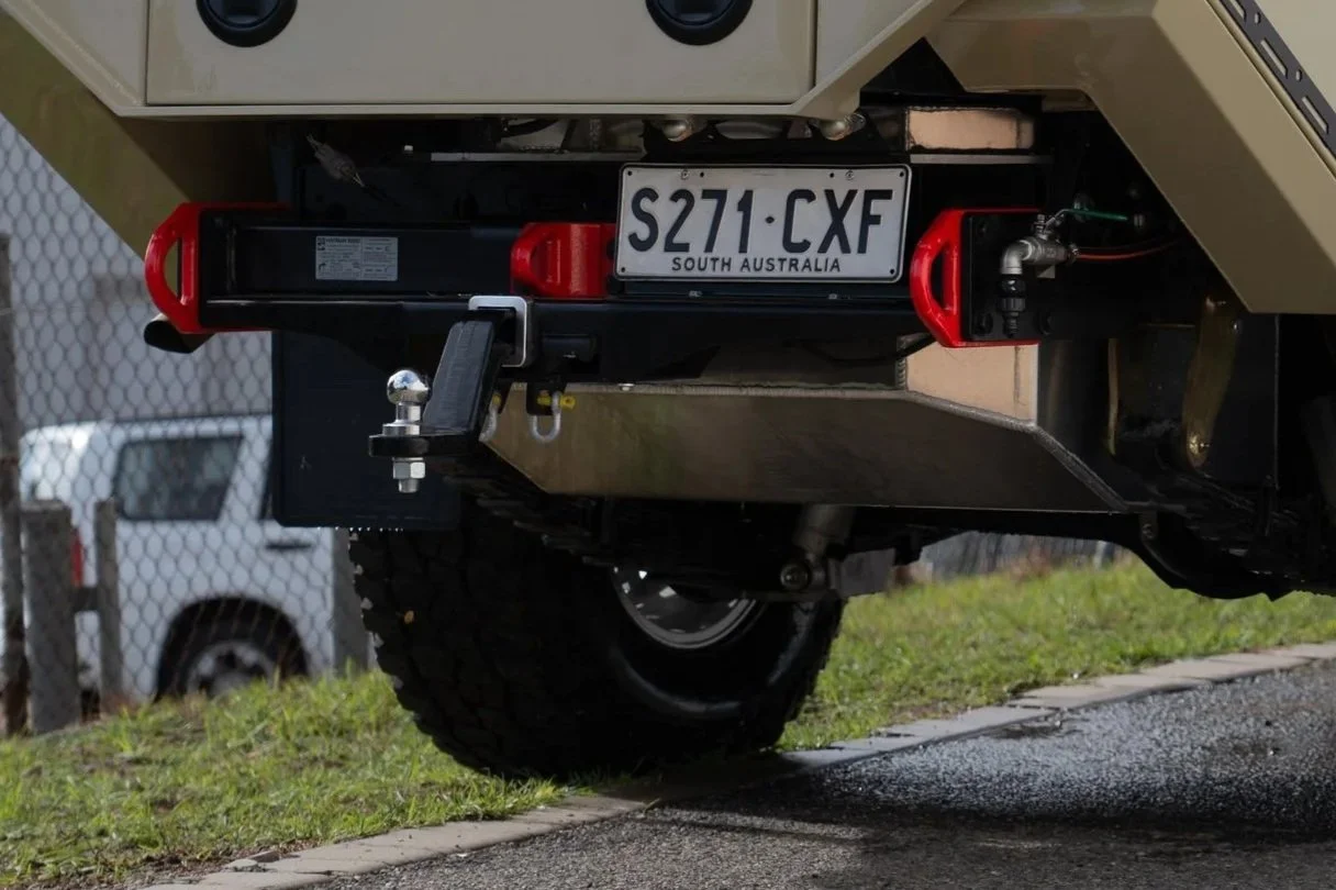 Close-up of the rear of a 79 series landcruiser, showing a custom fuel tank, a trailer hitch with a ball mount, and part of the vehicle's wheel and chassis, with a gravel driveway and a chain-link fence in the background.