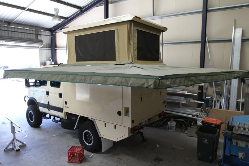 A beige camper truck with an extended pop-up roof and tent fabric. The camper is parked inside a garage with various tools and equipment around.