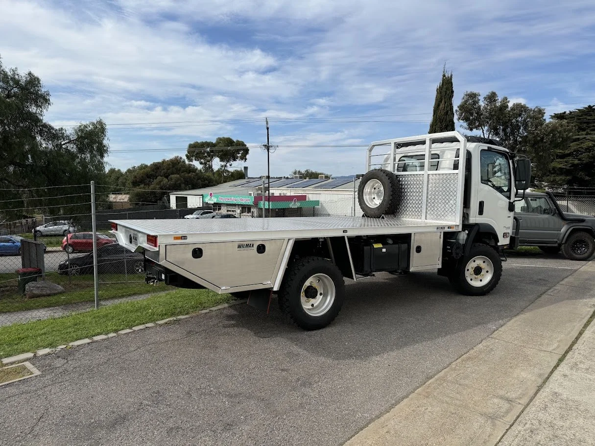 White flatbed truck parked on the side of the road with a chain-link fence behind it, a parking lot with several cars, and a building with a sign in the background. The sky is partly cloudy.