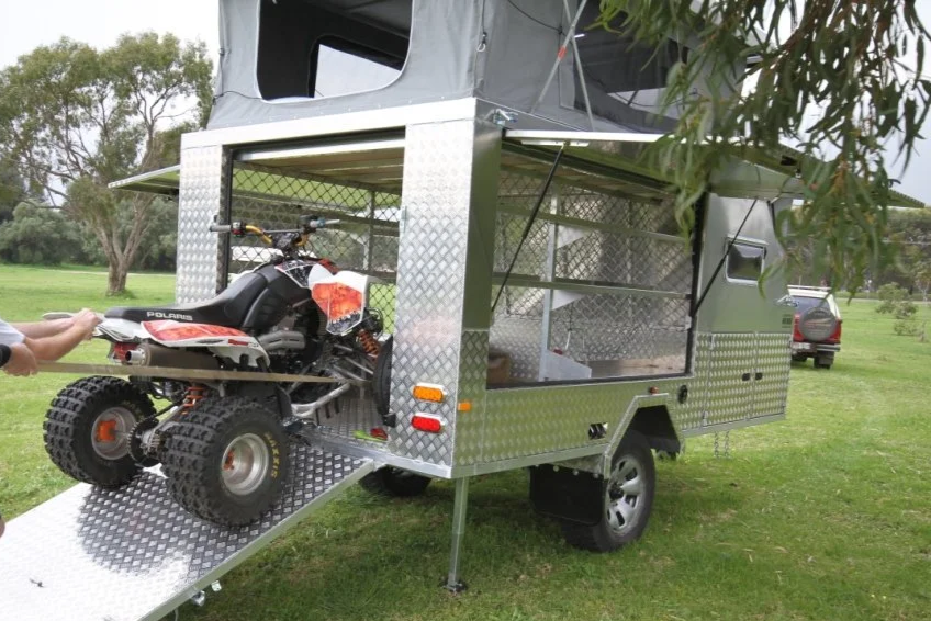 A custom toy hauler trailer, parked on a grassy area, with a dirt bike loaded on a lift at the front of the trailer.