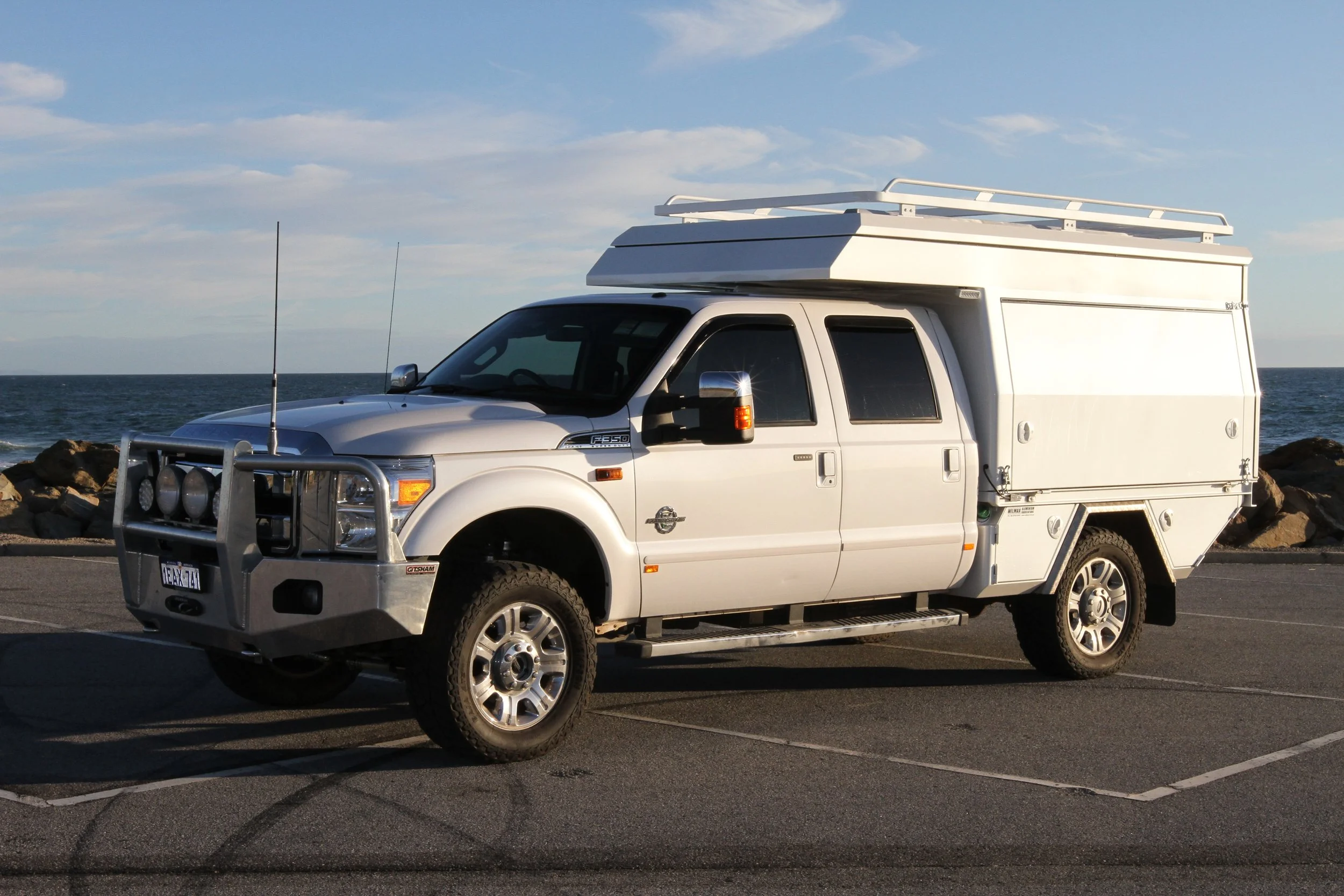 White utility truck parked near the ocean with rocky shoreline in the background, under partly cloudy sky. with custom pop top camper
