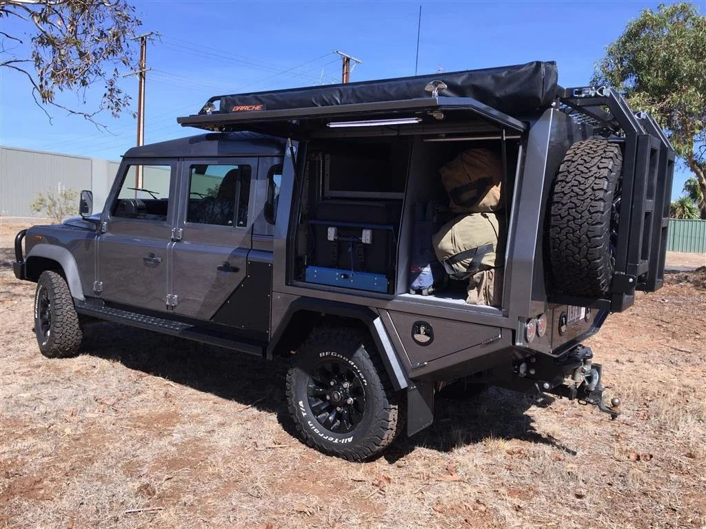 Black off-road vehicle with open rear storage area containing gear and supplies, parked on dirt ground with trees and power lines in the background.