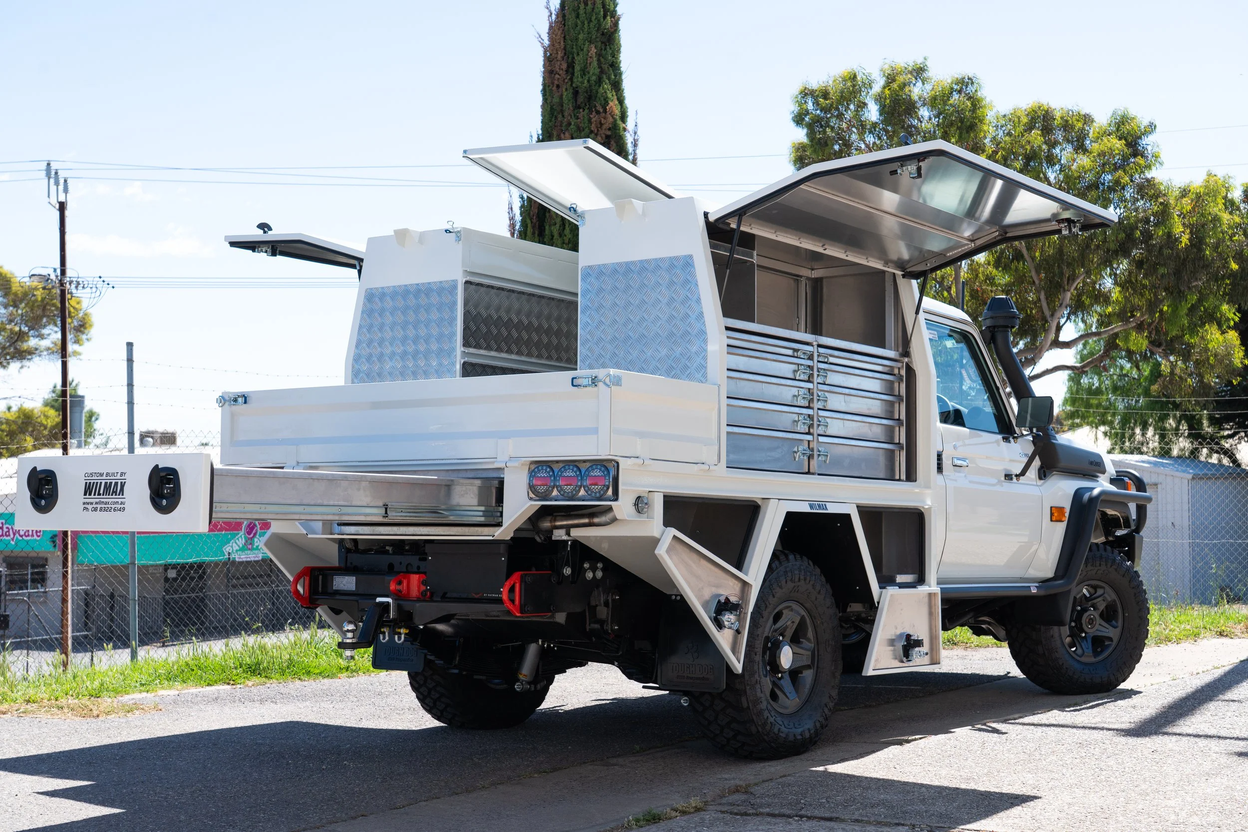 A white utility truck with open toolboxes and compartments, parked outdoors with trees and utility poles in the background.