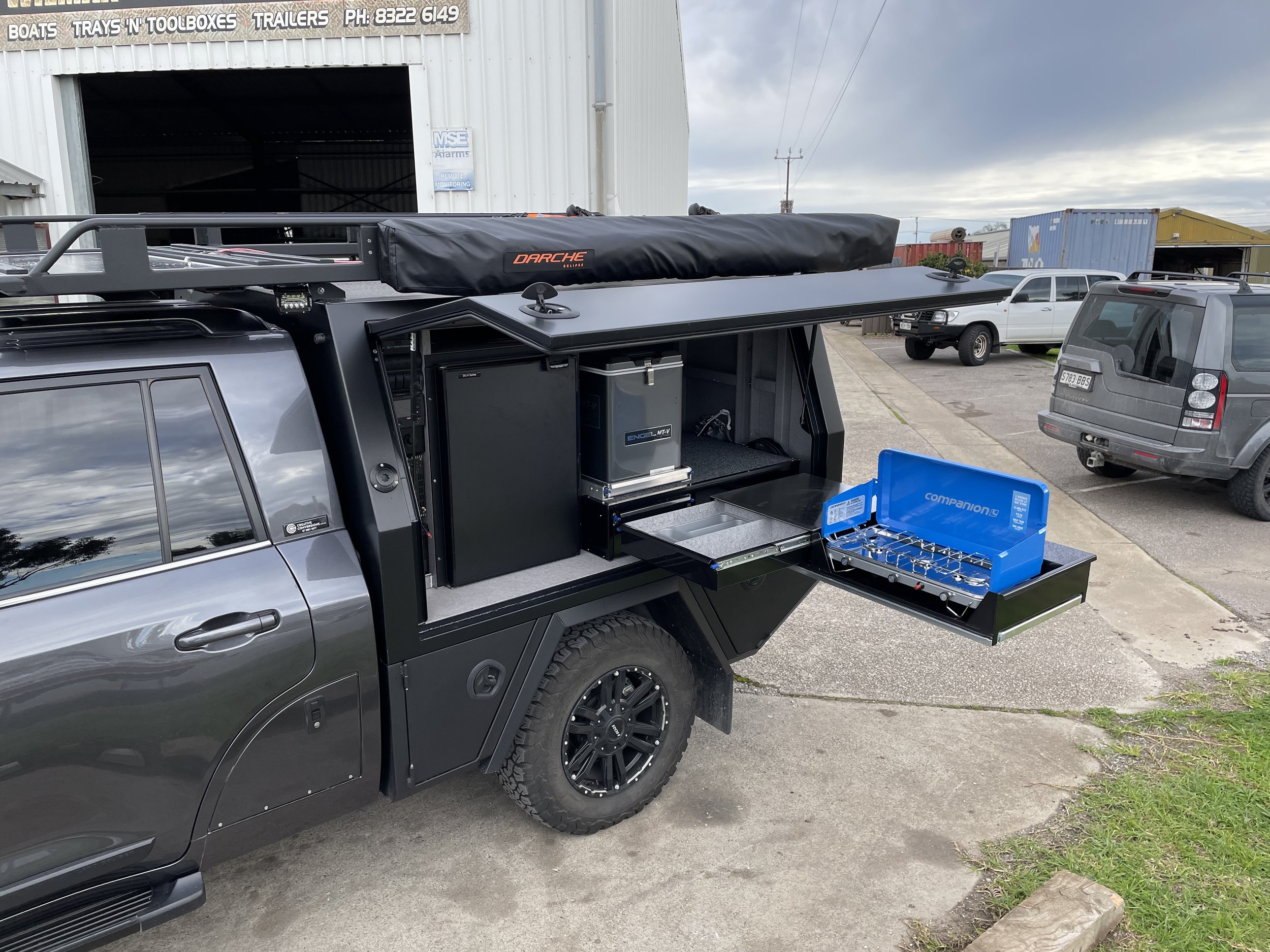 The image shows a gray SUV with its roof open, featuring a black roof rack with an orange logo, and open compartments with tools and equipment inside. The background includes a workshop or storage building with a sign that reads 'BOATS TRAYS 'N' TOOLBOXES TRAILERS' and a phone number. Several other vehicles are parked nearby, and the scene appears to be in an industrial or commercial area.