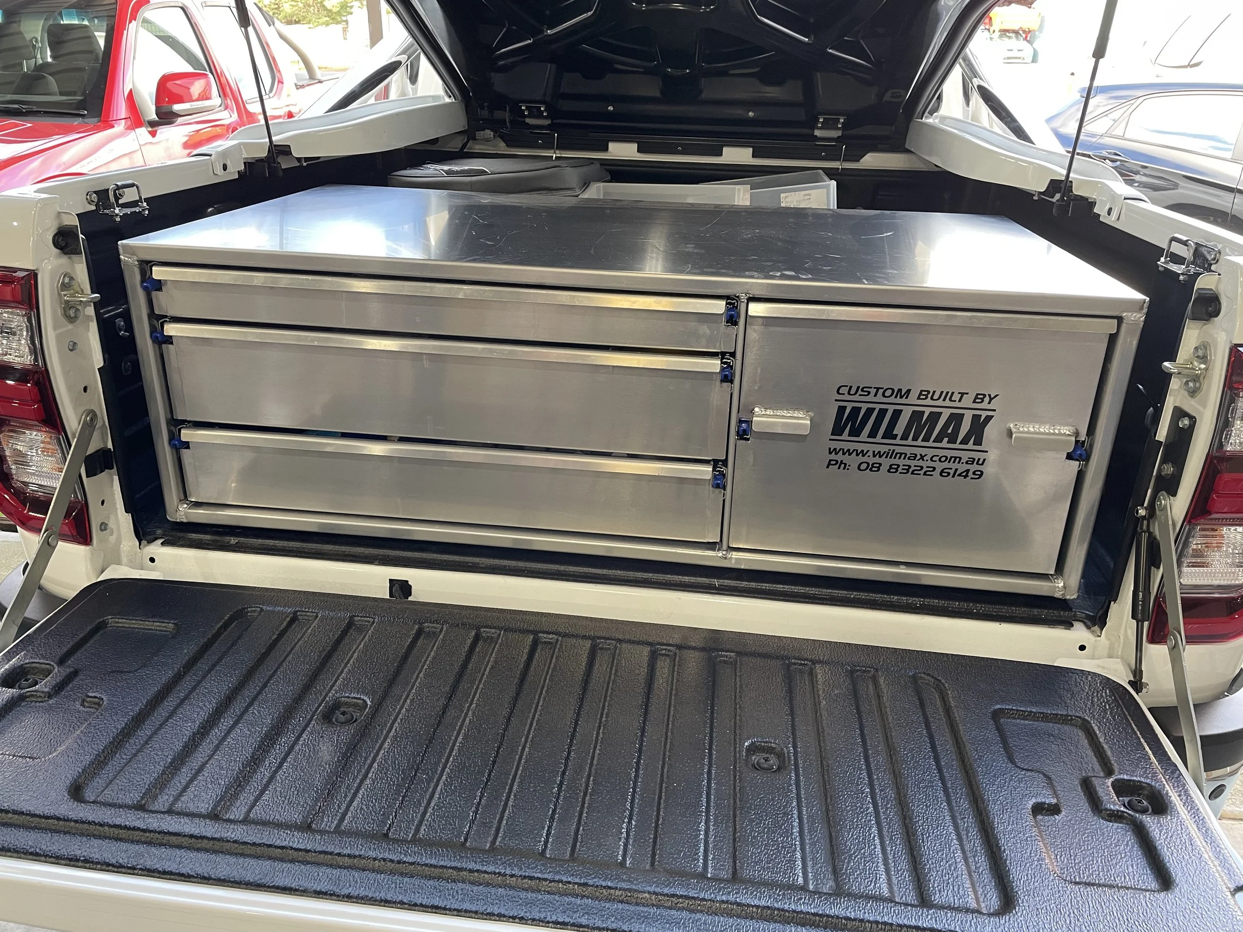 The image shows the open back of a white vehicle with a custom Wilmax aluminum storage system installed in the trunk area. The storage system features drawers and compartments, and the trunk floor is lined with a textured black mat.