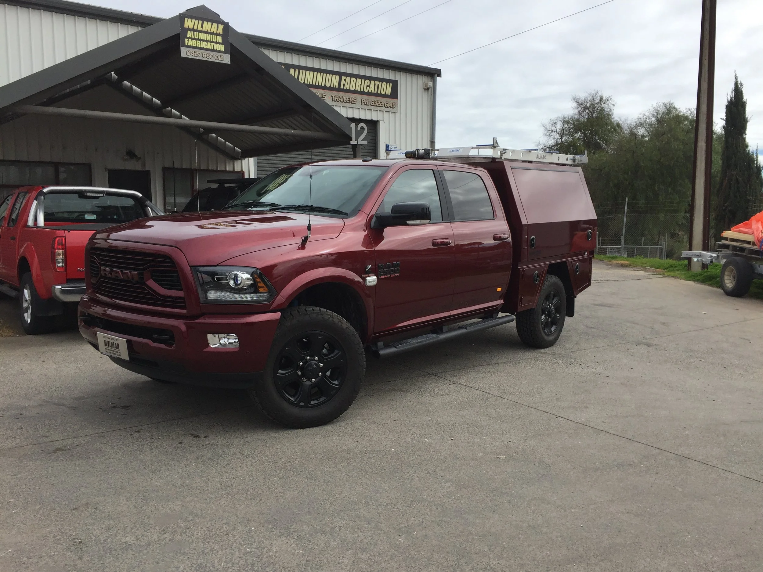 A maroon pickup truck with a custom tray and ladder on top parked in front of an aluminum fabrication shop. The shop has a gray metal exterior and signs advertising aluminum fabrication services.