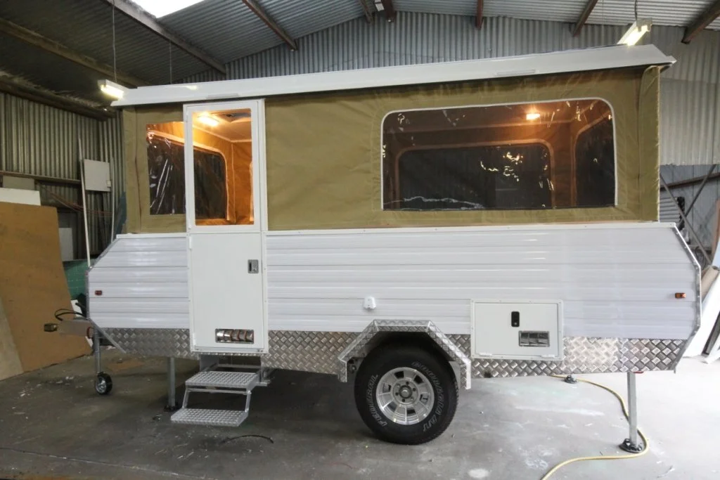 A white pop top camper trailer with a beige fabric cover and large windows, parked inside a warehouse.