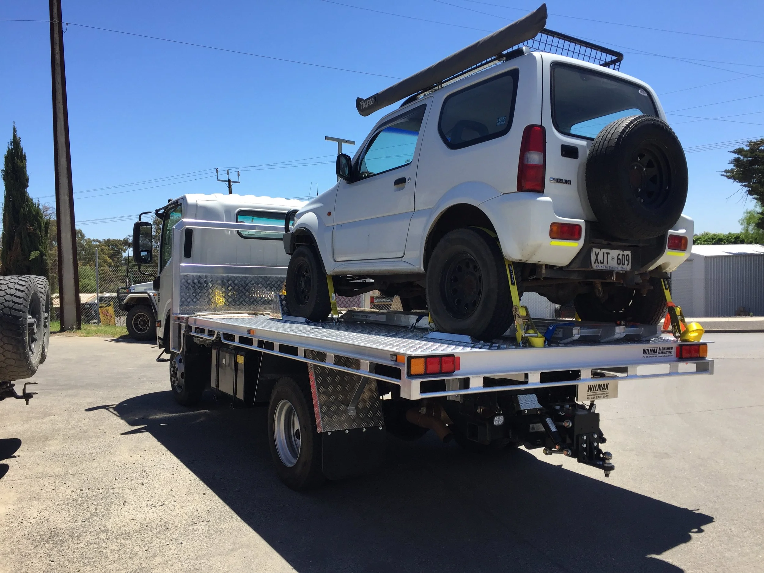 A white Suzuki vehicle on a flatbed tow truck, with a spare tire mounted at the back, parked outdoors on a sunny day with clear blue skies, utility poles, and trees in the background.