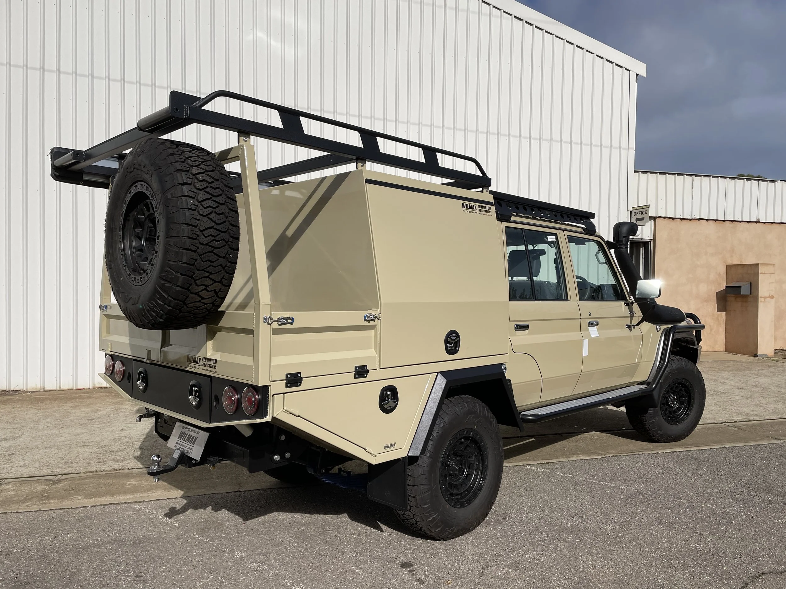 A beige 79 series with a custom canopy dog box with a spare tire mounted on the back, parked in front of a white industrial building with a small office sign. The vehicle has a black roof rack and is equipped for outdoor adventures.