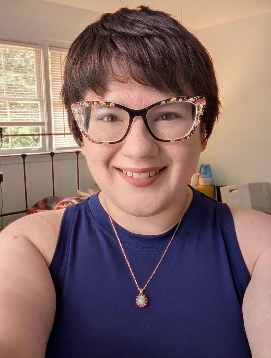 A person with short brown hair, wearing glasses with floral-patterned frames, a nose ring, and a necklace with a colorful pendant. They are smiling and wearing a dark blue sleeveless top. The background shows a room with windows, blinds, and some furniture.