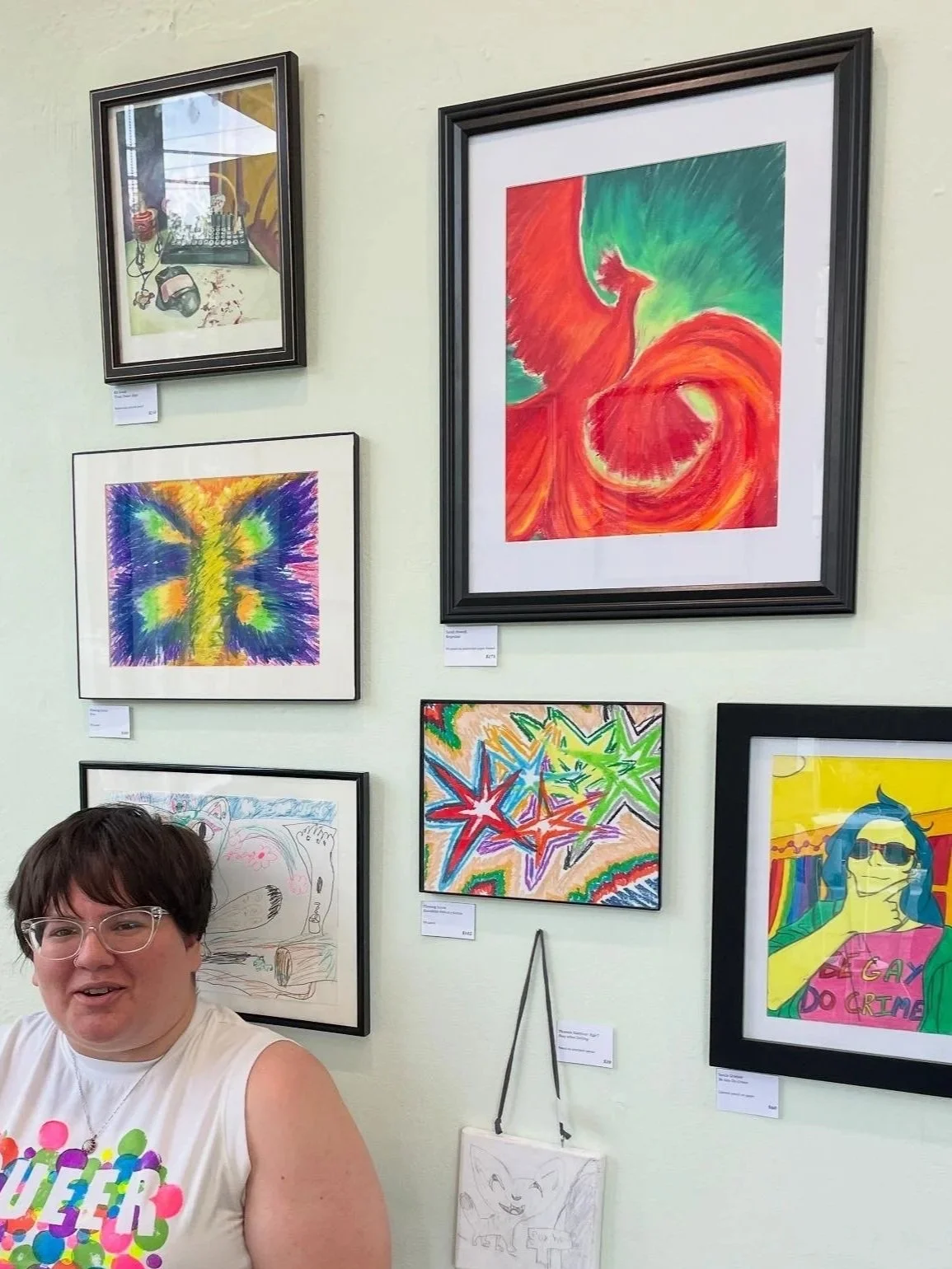 A person with short dark hair and glasses in a sleeveless shirt, smiling, standing in front of a wall with several framed colorful art pieces.