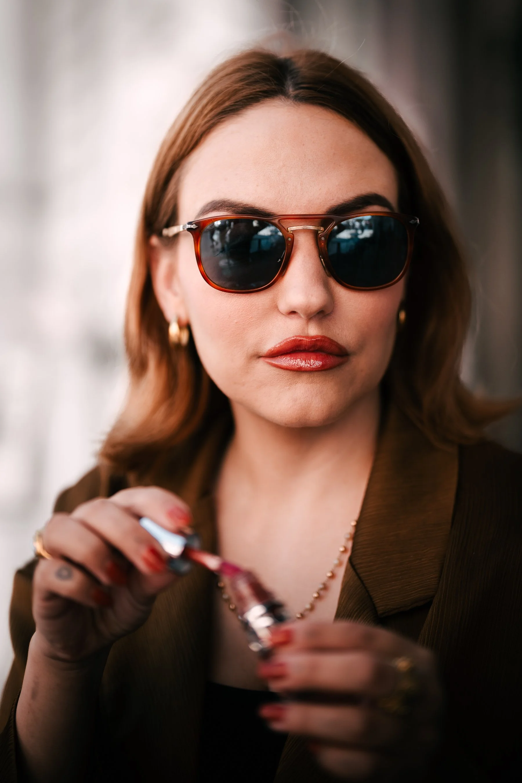 A woman with brown hair wearing sunglasses and gold earrings, holding a lipstick and applying it.