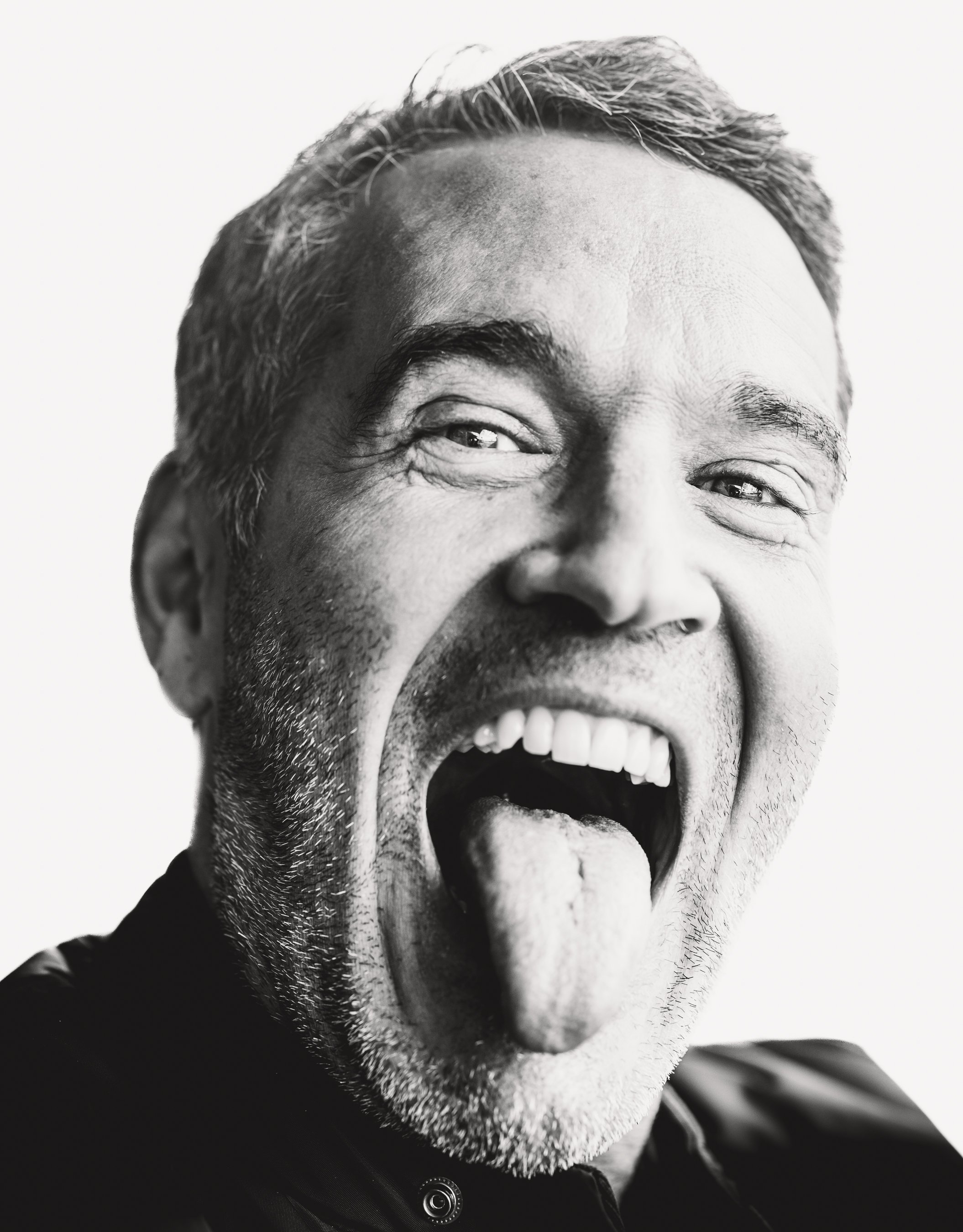 Close-up black and white photo of a smiling middle-aged man with salt-and-pepper hair, showing his tongue out.