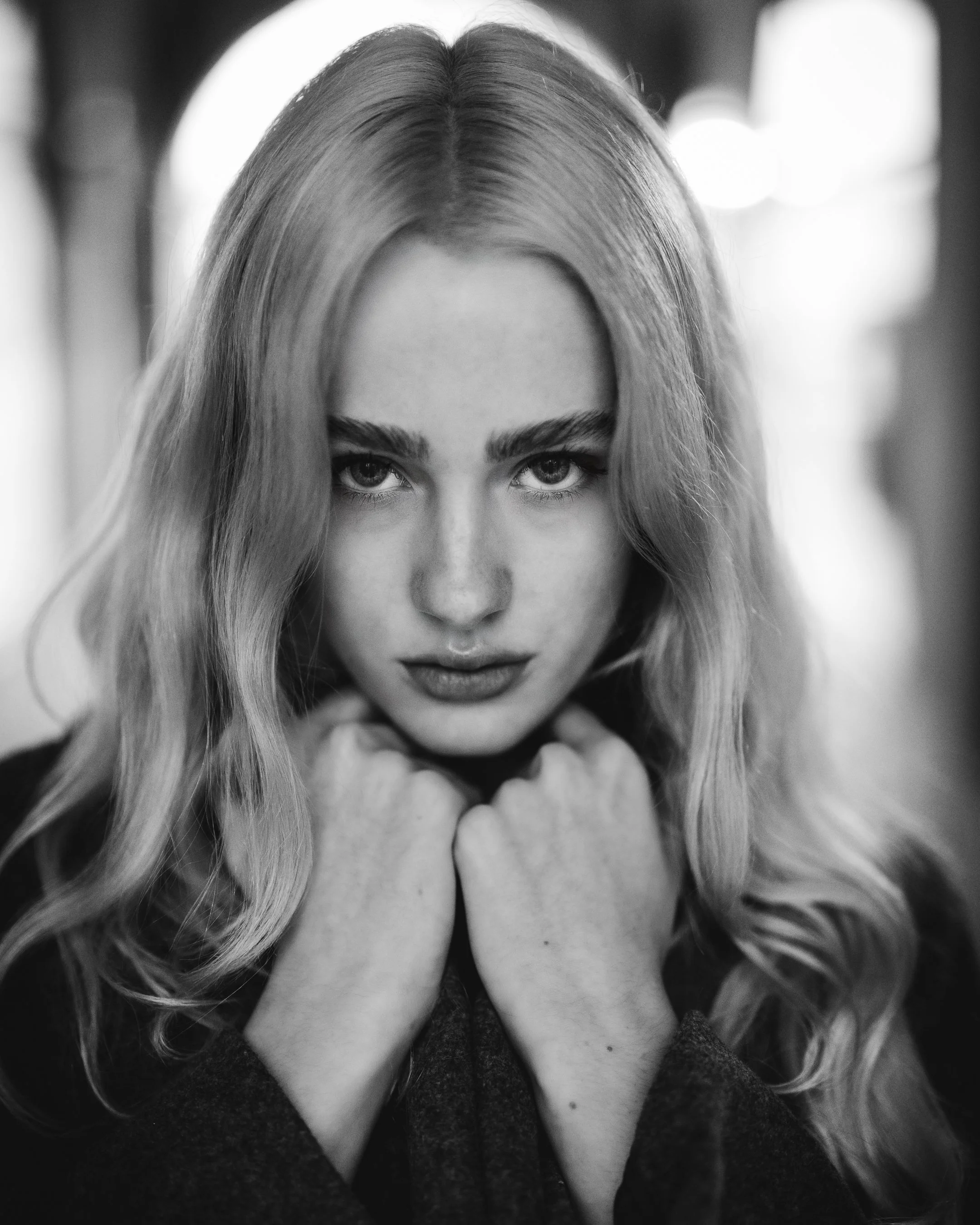 Black and white portrait of a woman with long, wavy hair looking directly at the camera, with her hands near her chin.
