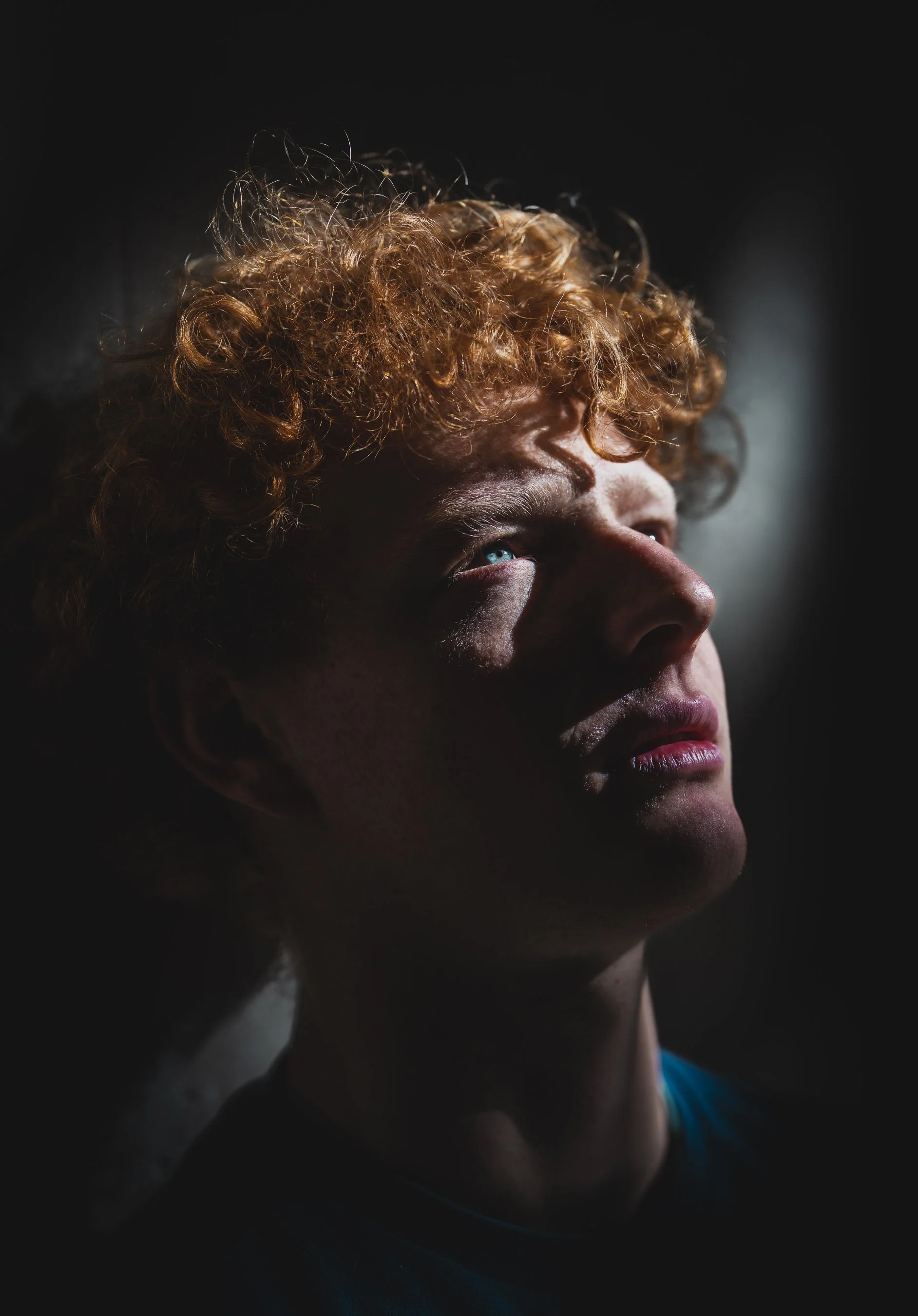 Close-up of a person's face with curly hair, illuminated from the side, showing detailed facial features against a dark background.