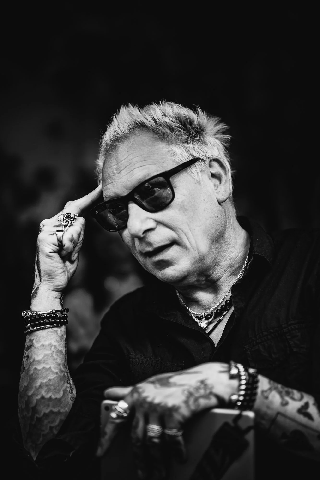 Black-and-white photo of a man with styled blonde hair wearing dark sunglasses, multiple rings, bracelets, and necklaces, sitting at a table, holding a phone, with a dark blurred background.