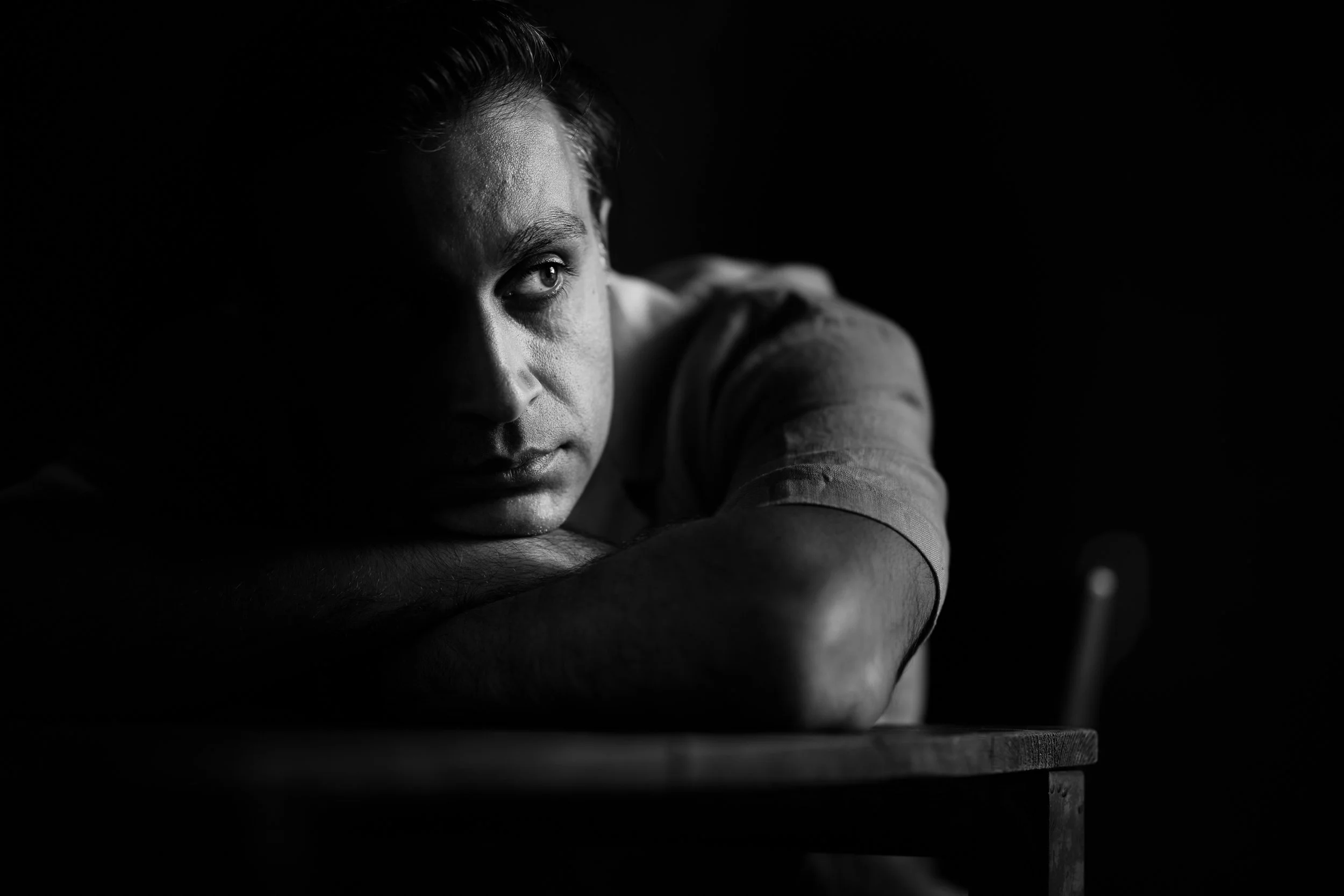 Black and white portrait of a man resting his head on his crossed arms on a table, looking thoughtfully to the side.
