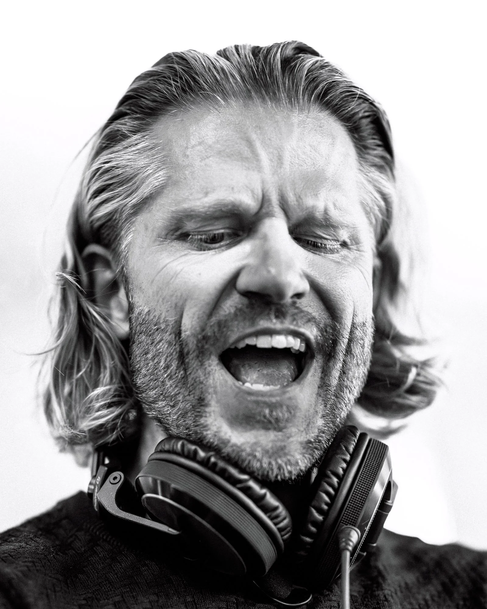 Black and white close-up photo of a man with long hair and a beard, wearing headphones around his neck, with his mouth open and eyes partially closed.