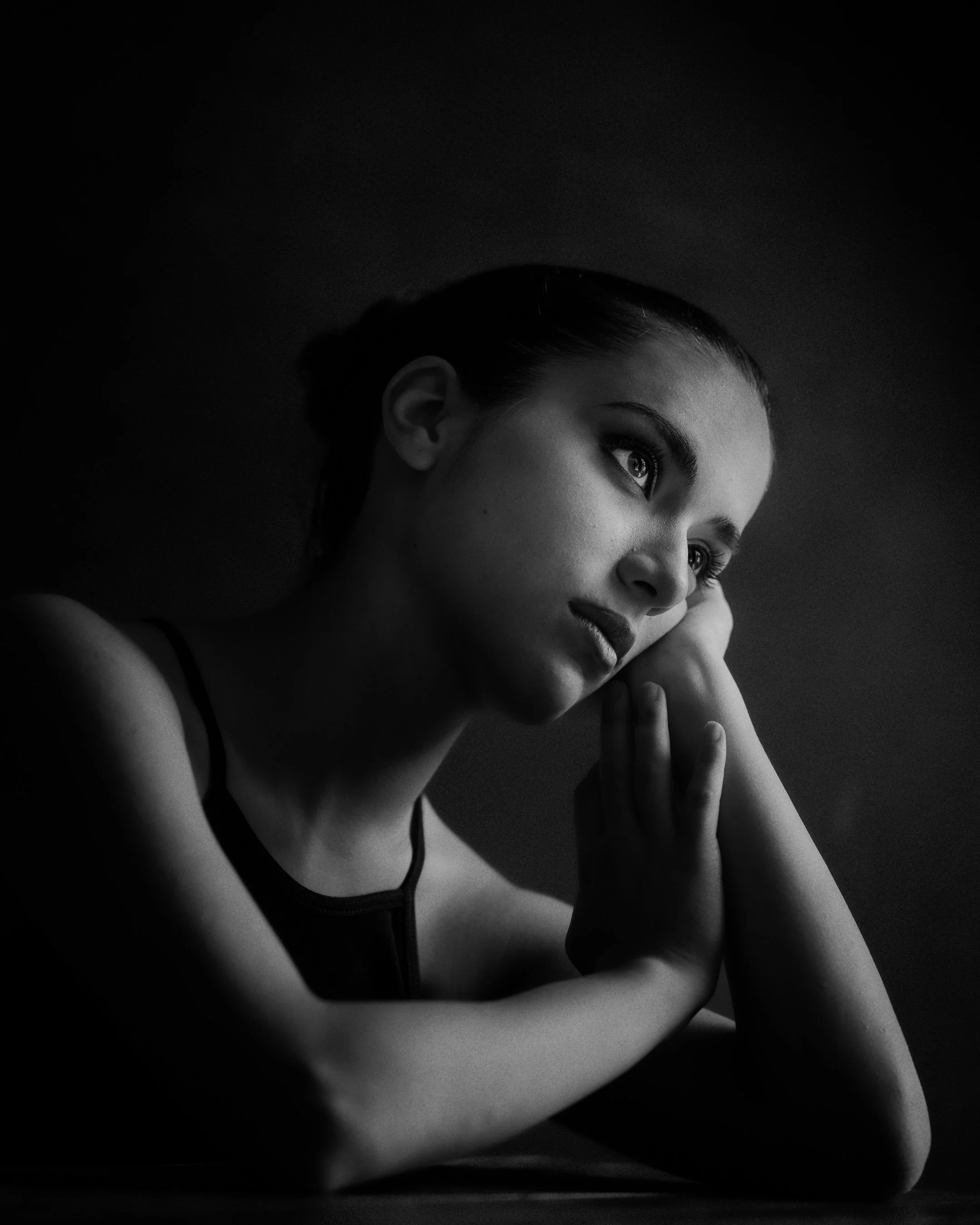 A black and white portrait of a young woman with her head resting on her hand, gazing thoughtfully to the side.