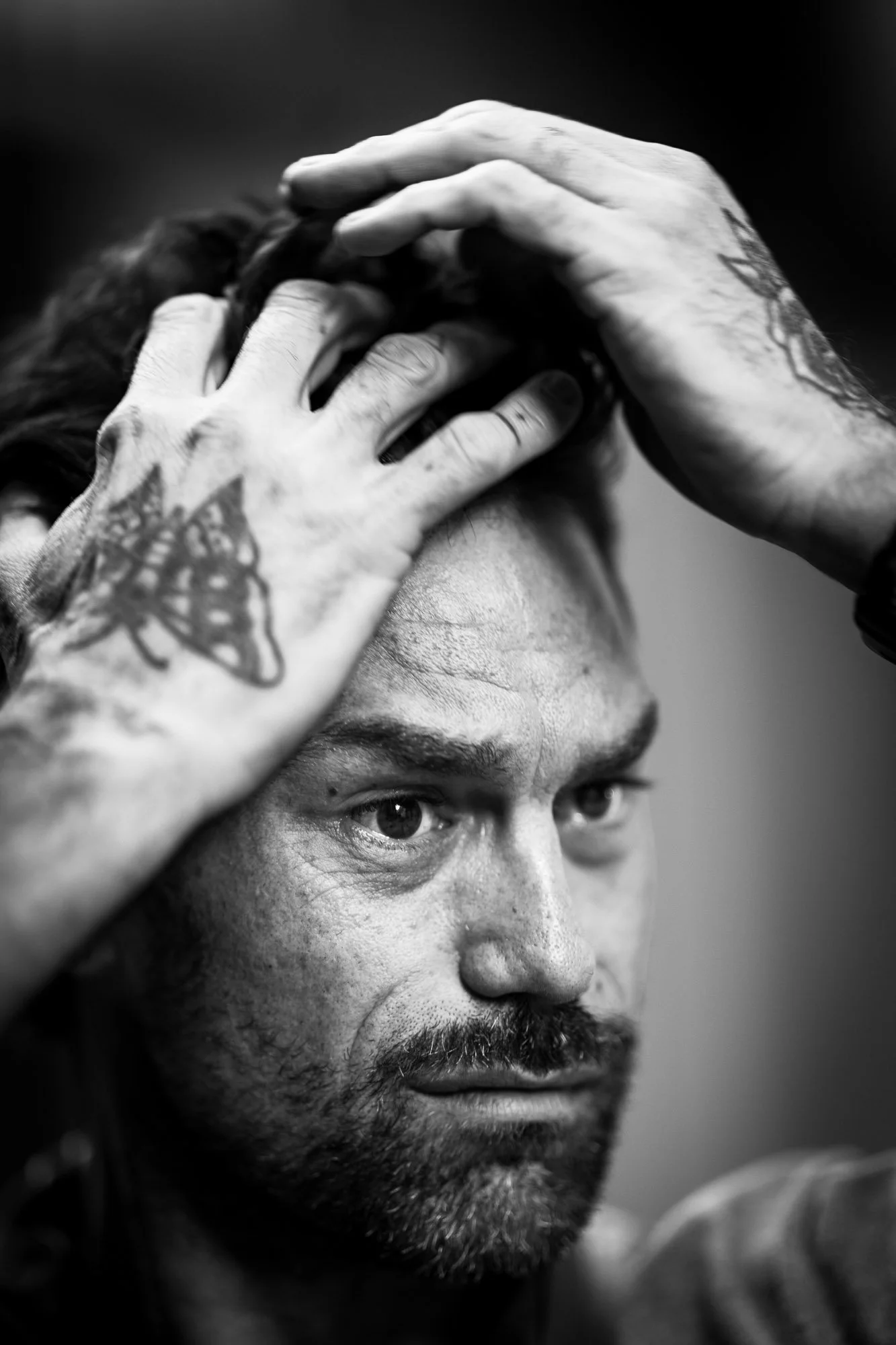 A black and white close-up photo of a man with tattoos on his hands and forehead, with intensely focused eyes, holding his hands on his head.