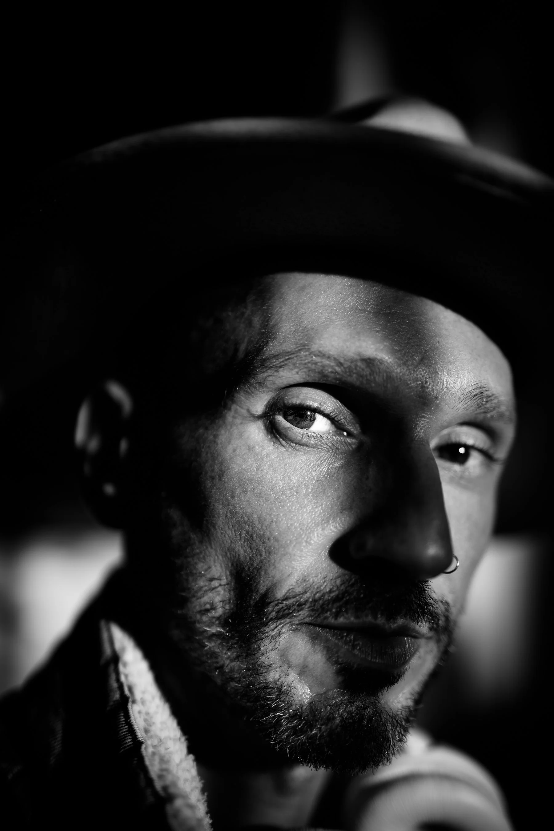Black and white close-up portrait of a man wearing a hat, with one eye slightly squinting and a nose piercing, looking directly at the camera.