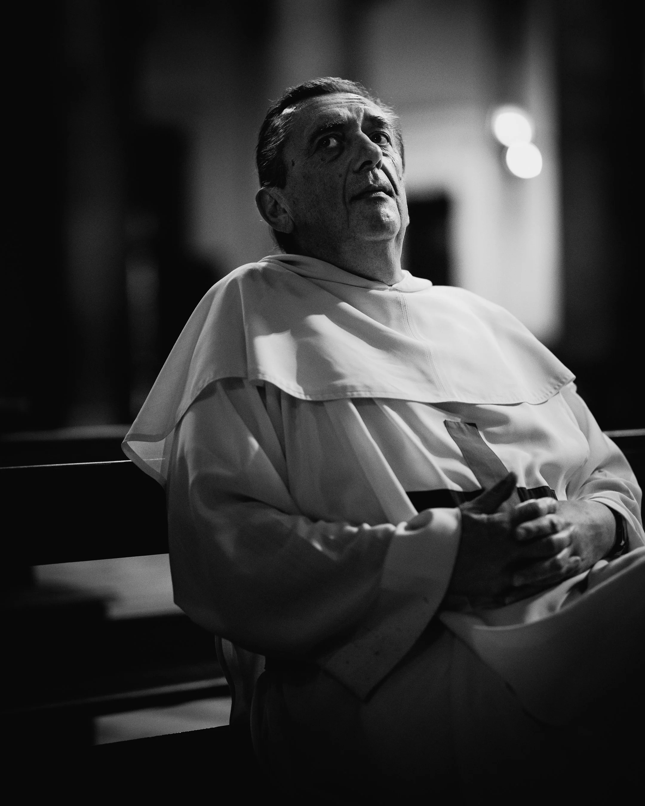 A black and white photo of a man in religious robes, sitting on a bench with his hands clasped, looking upward with a serious expression.