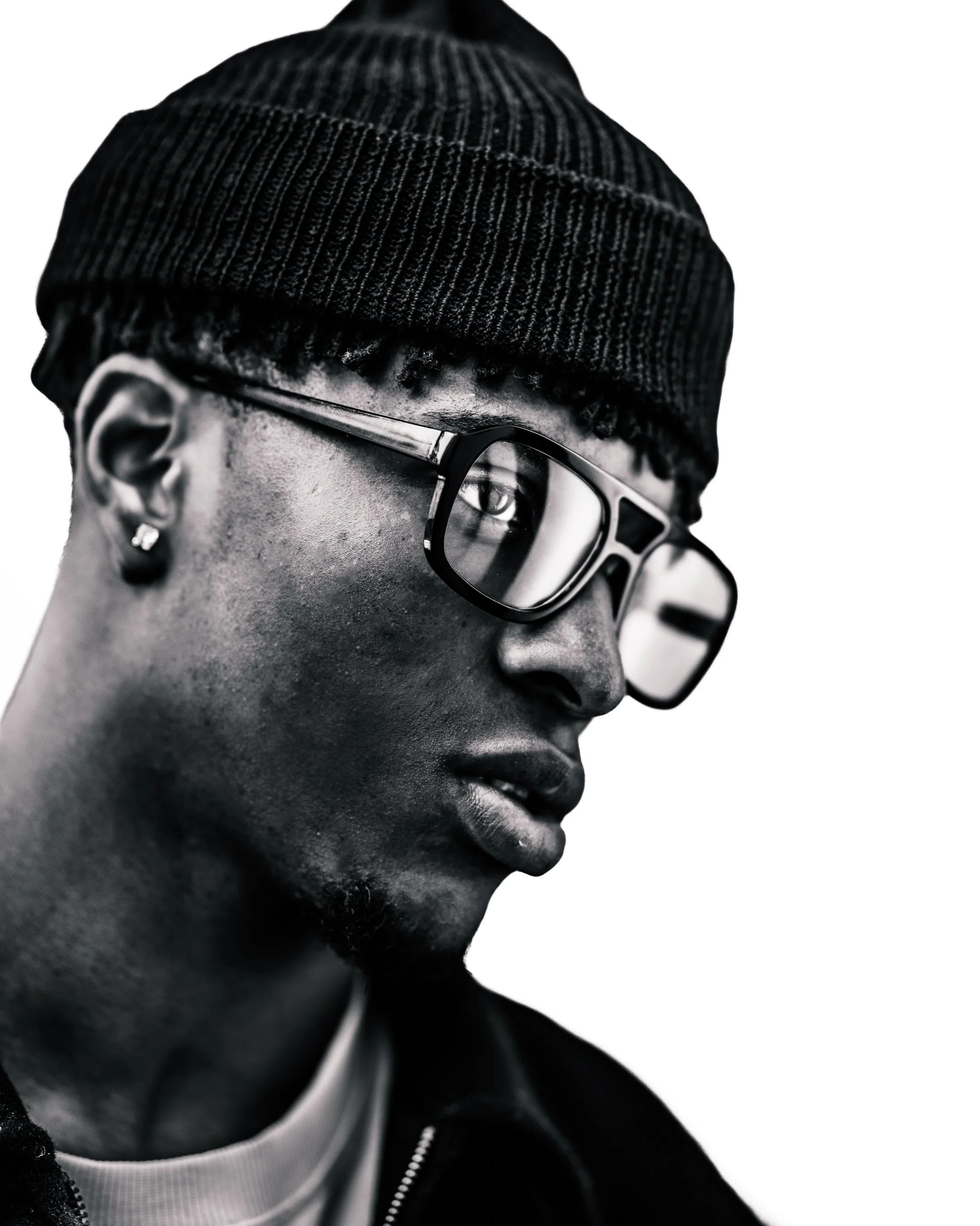 Black and white close-up portrait of a man with a knit cap, eyeglasses, and an earring, facing sideways.