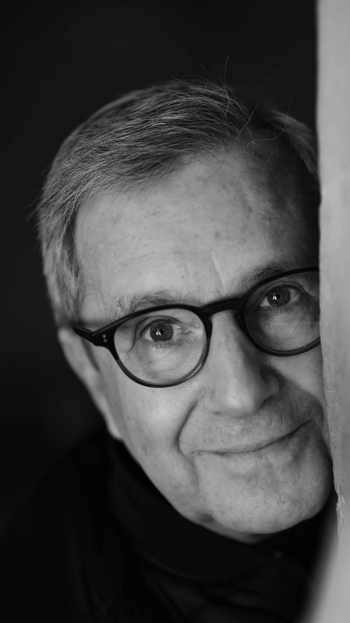 Close-up black and white photograph of a person with short gray hair, wearing round glasses, partially hidden behind a surface, smiling gently.