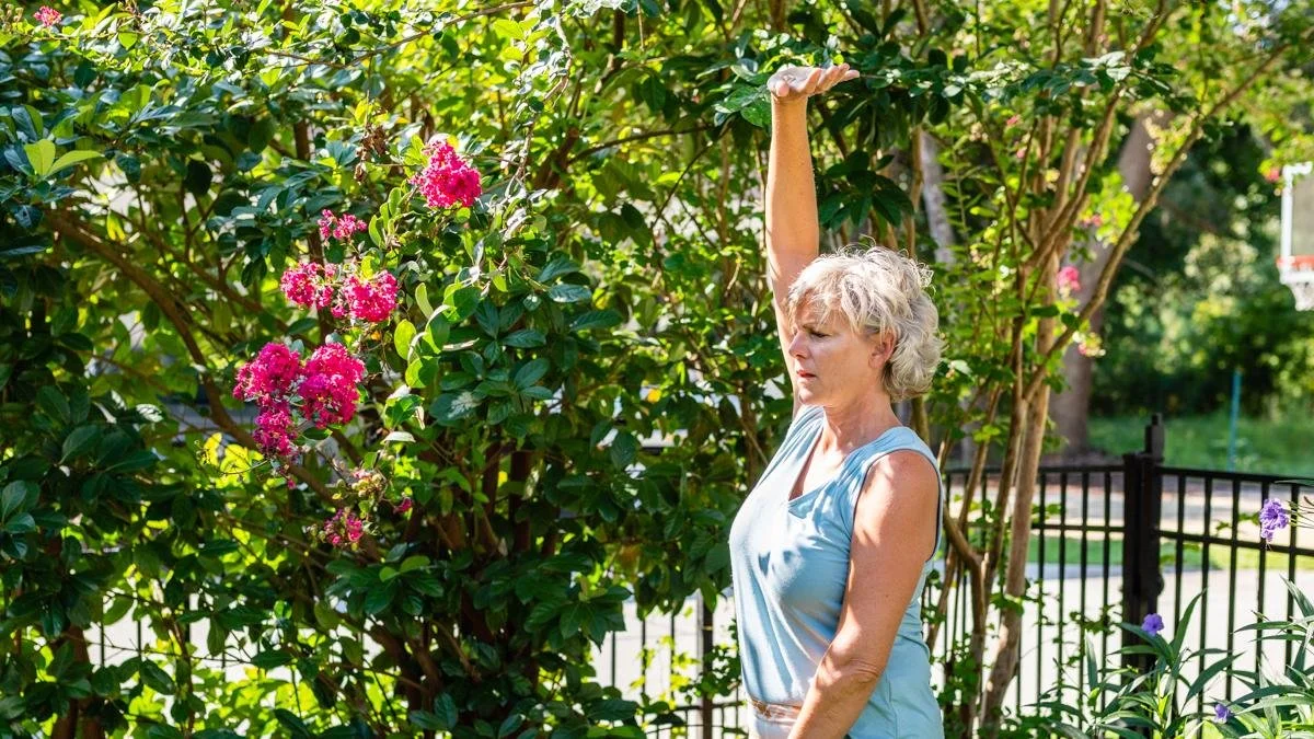 Woman practicing Qigong outdoors beside blooming pink crepe myrtle, moving her body as one whole person