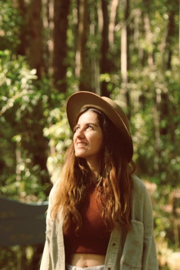Young woman with long wavy hair wearing a wide-brimmed hat and casual clothing, looking up in a forest.