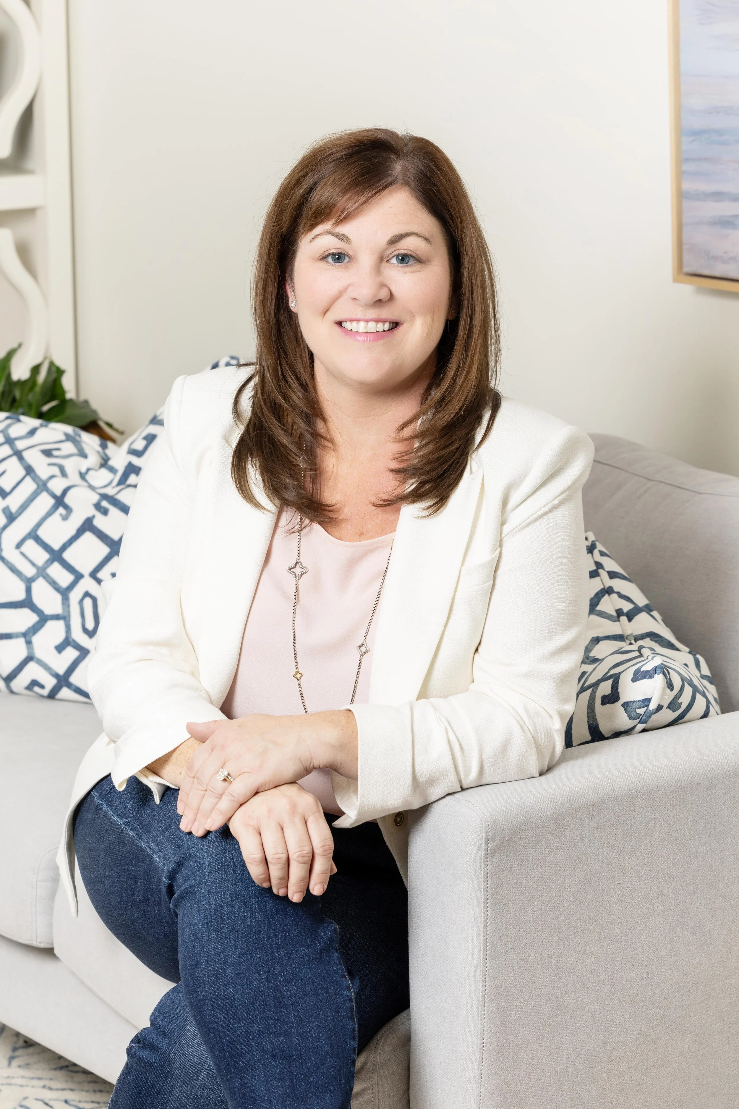 A woman with shoulder-length brown hair, wearing a white blazer and a light pink blouse, sitting on a beige sofa with blue and white patterned pillows, smiling at the camera in a cozy room with artwork on the wall and a plant nearby.