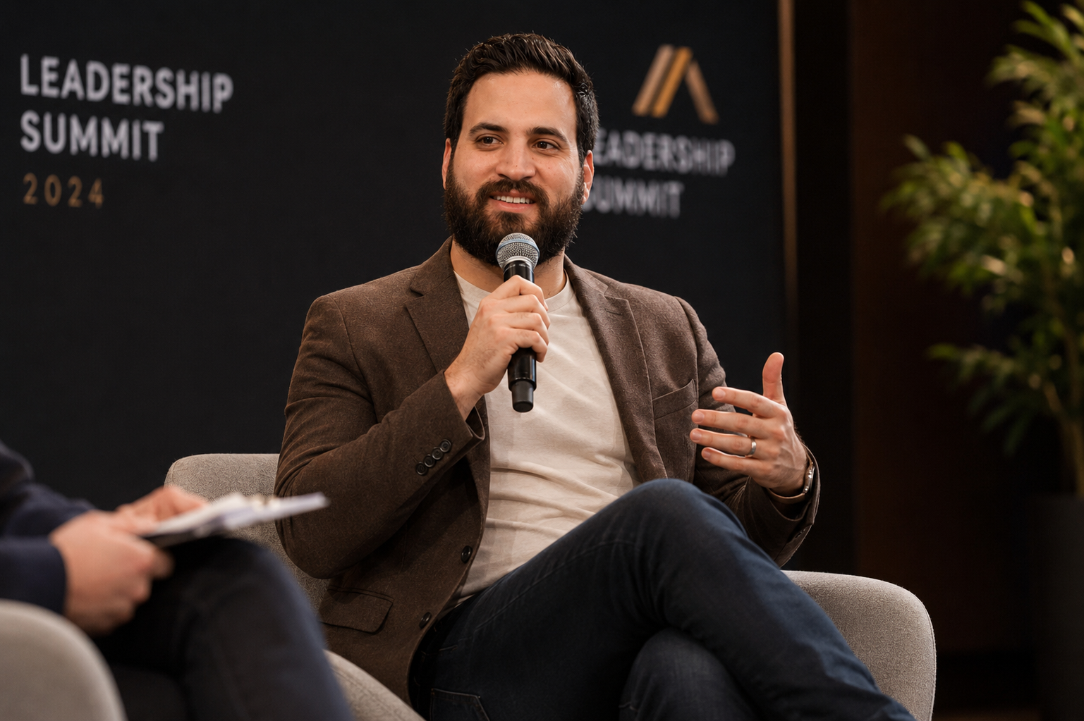 A man with a beard and dark hair speaking into a microphone at a leadership summit conference, seated on a gray chair, wearing a brown blazer and a white shirt.