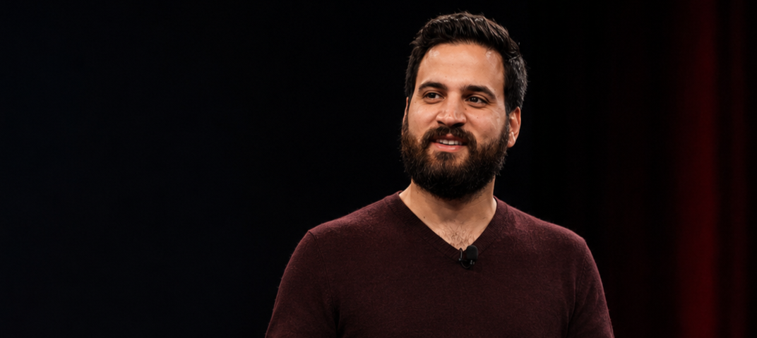 A man with dark hair and beard, wearing a maroon sweater, standing on a stage with a black and red background.