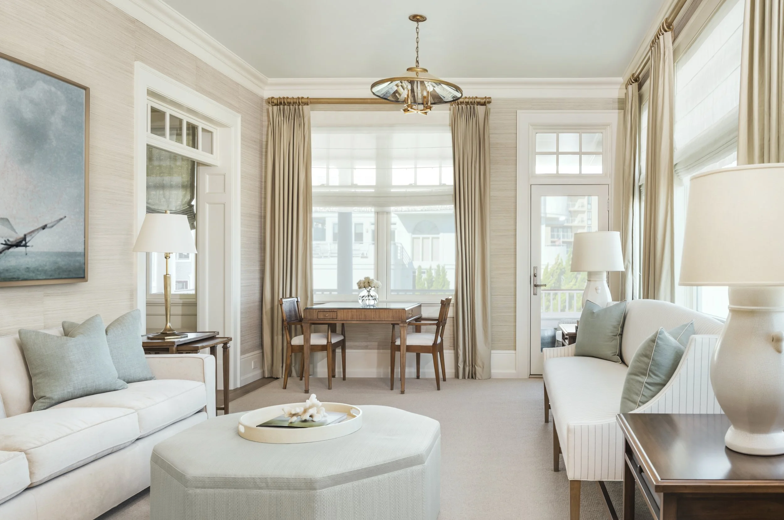 Bright living room with white sofas, a round ottoman, light-colored walls, large windows with beige curtains, a wooden dining table with chairs, and a modern ceiling light fixture.
