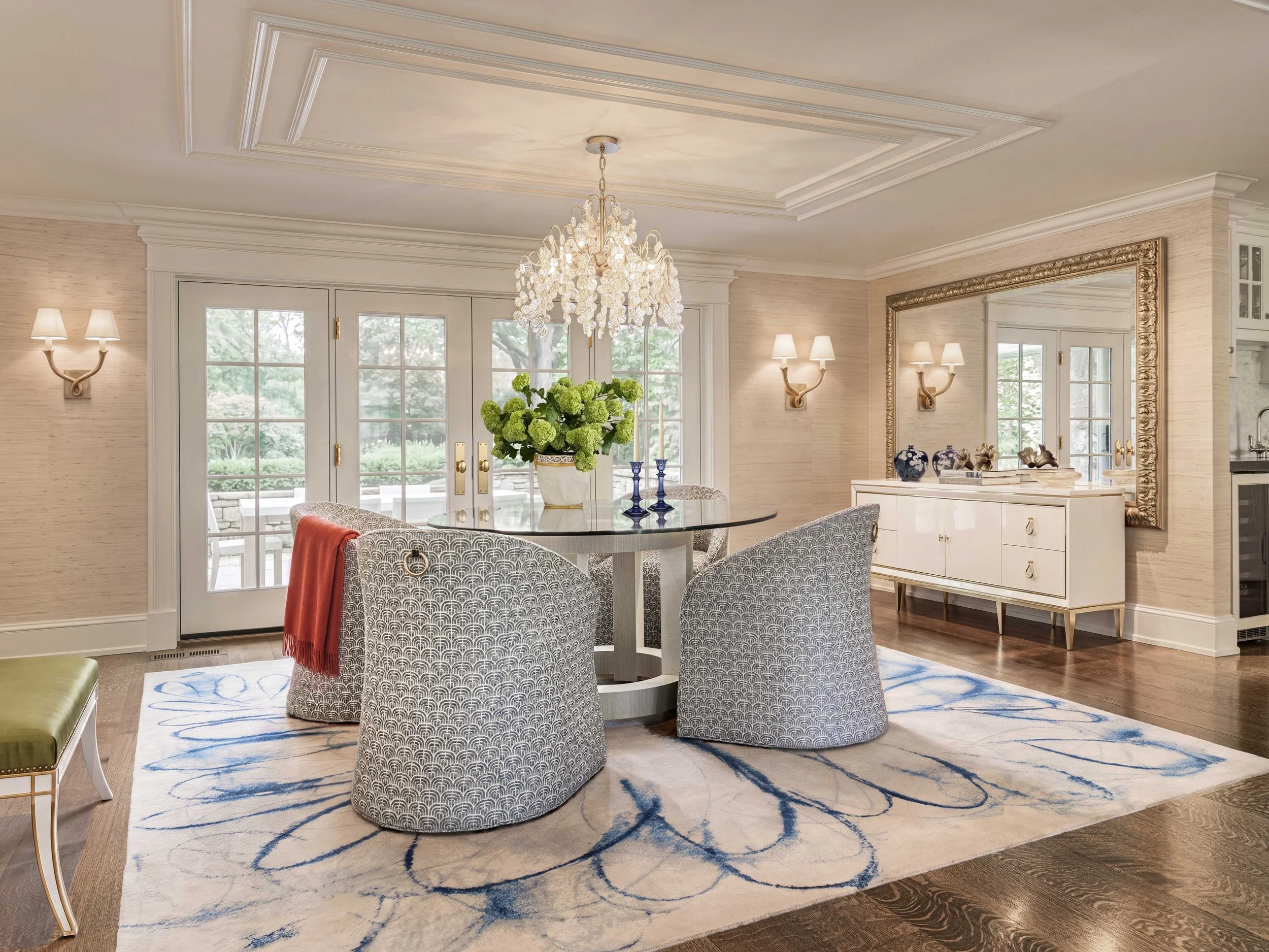 A spacious, elegantly decorated living room with a round glass dining table surrounded by four patterned chairs. A large chandelier hangs above the table, and a floral arrangement is in the center. The room features cream-colored walls, wall sconces, a large mirror with an ornate gold frame, and French doors leading to an outdoor patio visible through the glass. There are decorative items on a sideboard, including blue vases and sculptures. An area rug with a blue and white pattern covers part of the wooden floor.